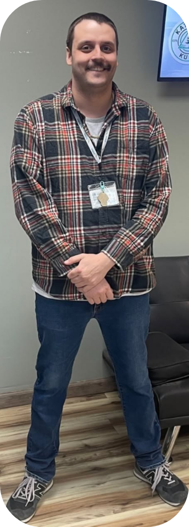 Young man with a beard and mustache, wearing a plaid shirt and jeans, standing indoors with a lanyard and badge, smiling, with a television screen in the background. Web design