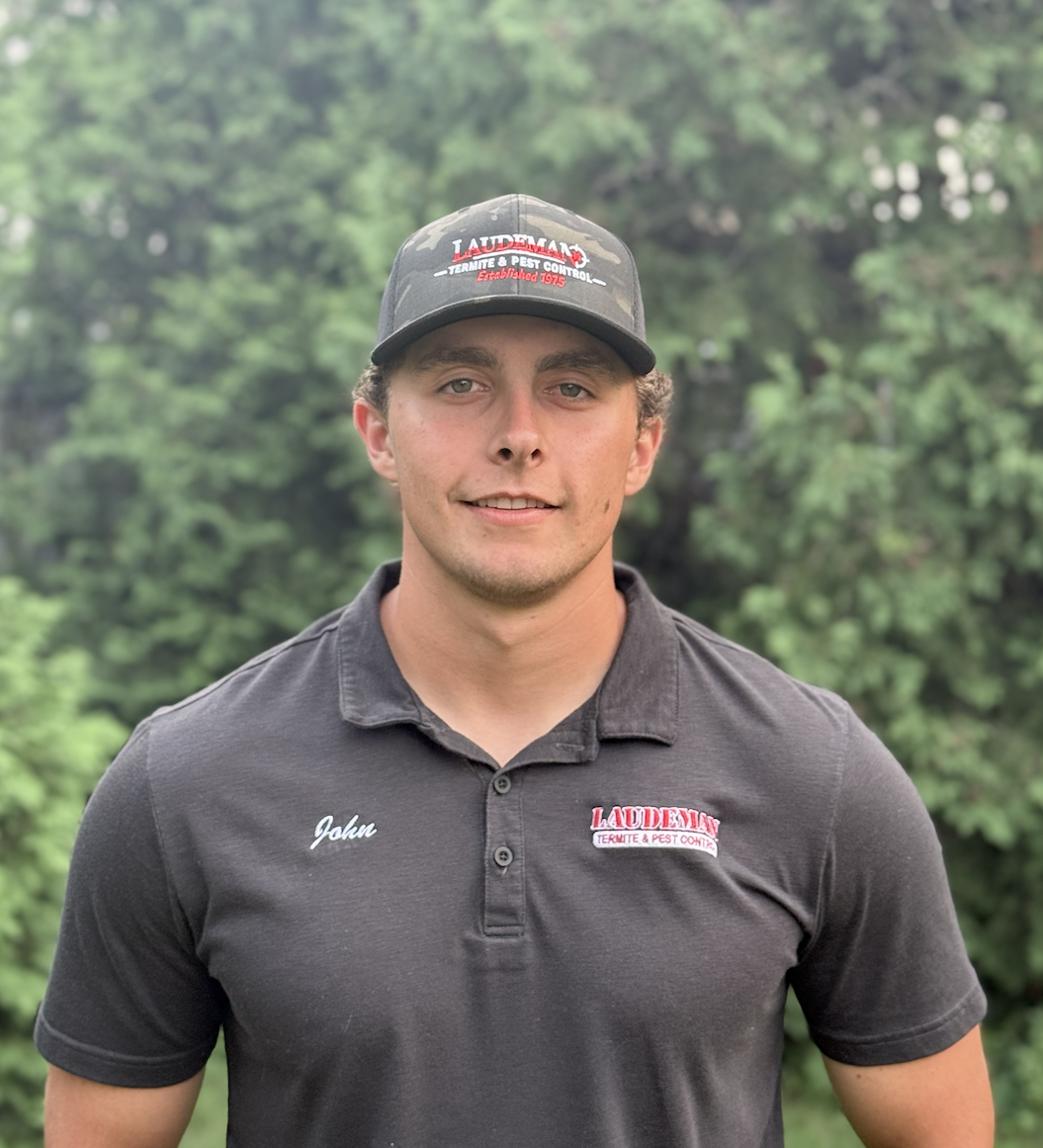 A young man wearing a black company uniform with 'John' embroidered on the left chest and a black cap with the 'Laudeman Termite & Pest Control' logo, standing outdoors with a blurred green trees background.