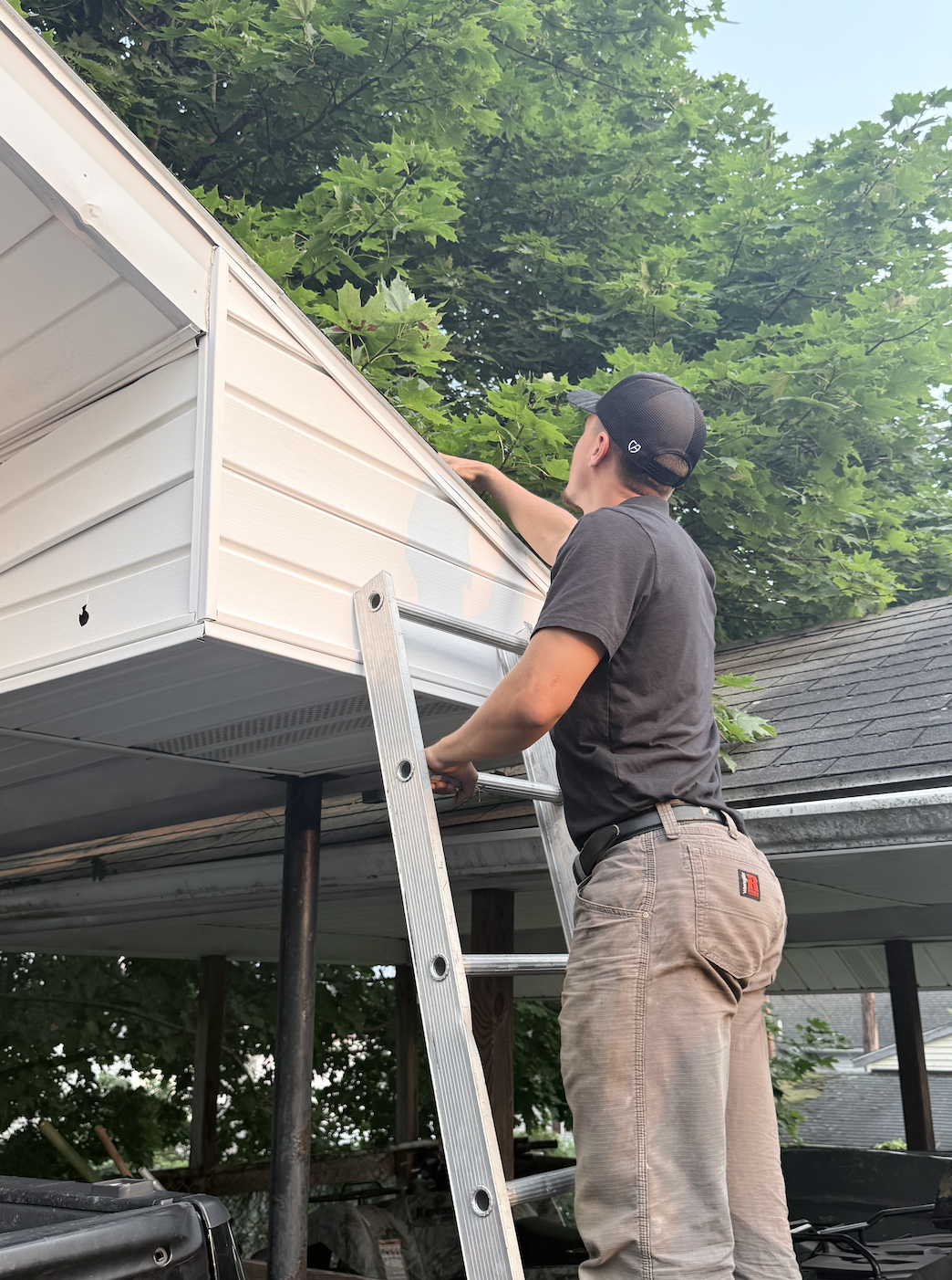 A man wearing a black cap and gray t-shirt standing on a ladder, working on the gutter of a house with beige siding, surrounded by green trees.