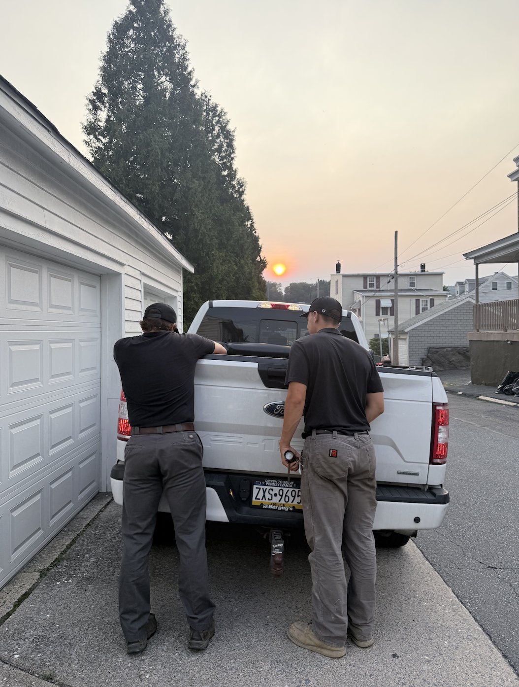 Two men standing at the back of a white pickup truck parked in a driveway during sunset. One is leaning on the truck's tailgate, and both are wearing black shirts and gray pants, with one holding a flashlight. Residential houses and a large tree are visible in the background.