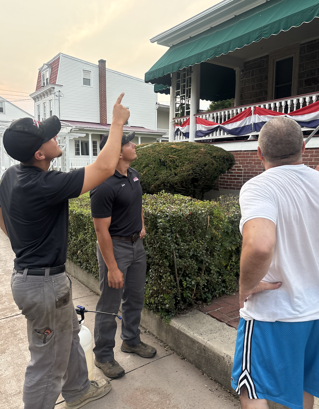Three men are standing outside a house with an American flag bunting on the porch. One man is pointing up at the house while the other two look on, with one holding a sprayer.