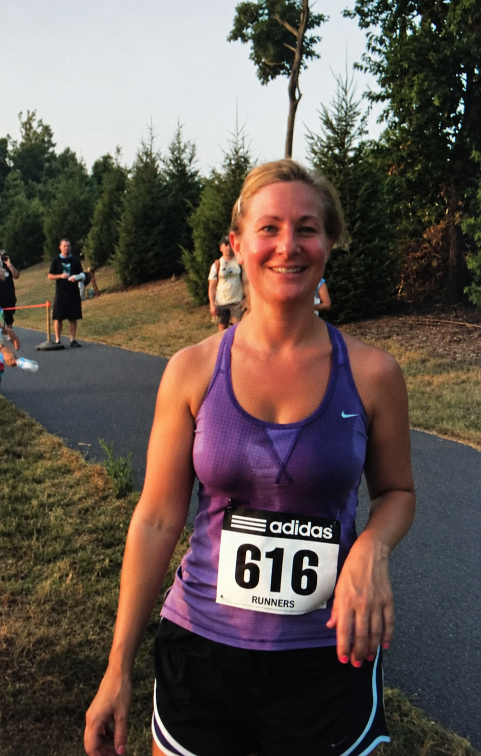 Smiling female runner wearing a purple Nike tank top and black shorts, bib number 616, during a race outdoors with trees and other runners in the background.