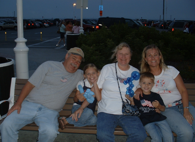 Family sitting on a park bench at dusk, with a parking lot in the background, smiling and holding balloon animals.