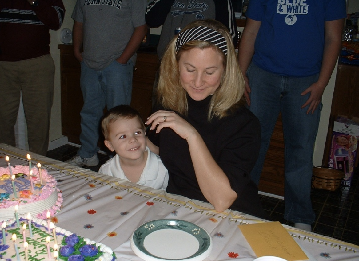 A woman and a young boy celebrating a birthday with a decorated cake with lit candles, surrounded by friends or family, at a table covered with a white tablecloth and floral decorations.