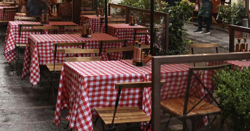 Outdoor restaurant seating with tables covered in red and white checkered tablecloths, wooden chairs, and condiment bottles on the tables.