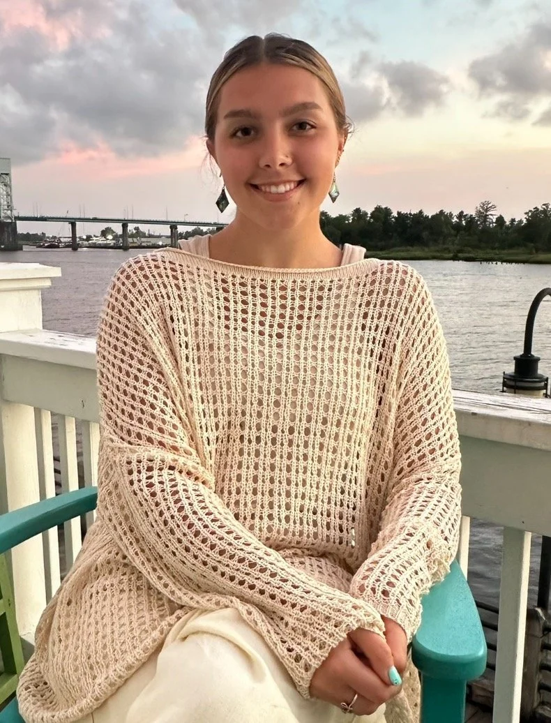 A young woman with blonde hair and earrings sitting on a porch near a body of water during sunset, wearing a cream-colored crocheted sweater.