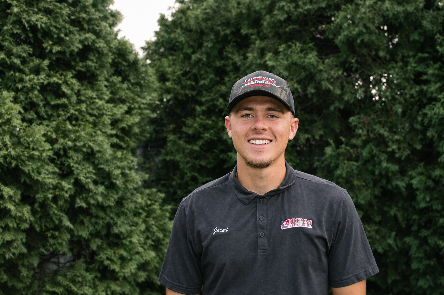 A young man smiling outdoors, wearing a black uniform with a name tag 'Jared' and a cap with 'LADLEMAN Termite & Pest Control' logo, standing in front of green foliage.