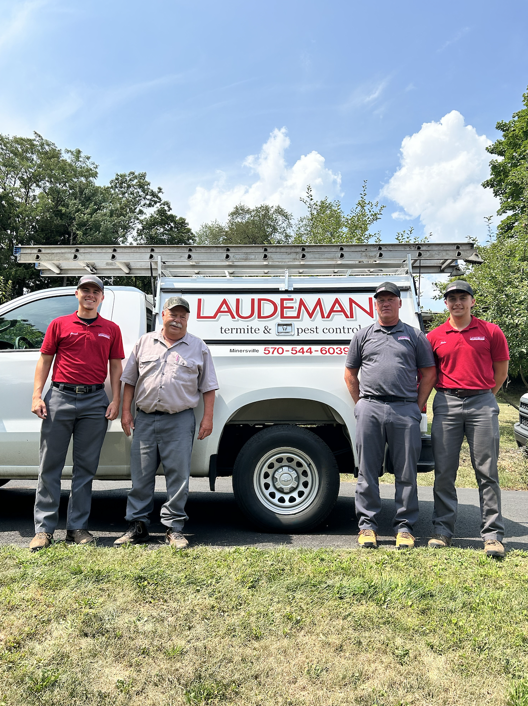 Four workers standing in front of a white pest control service truck with ladders on top, wearing red and gray uniforms, outdoors with trees and sky in the background.