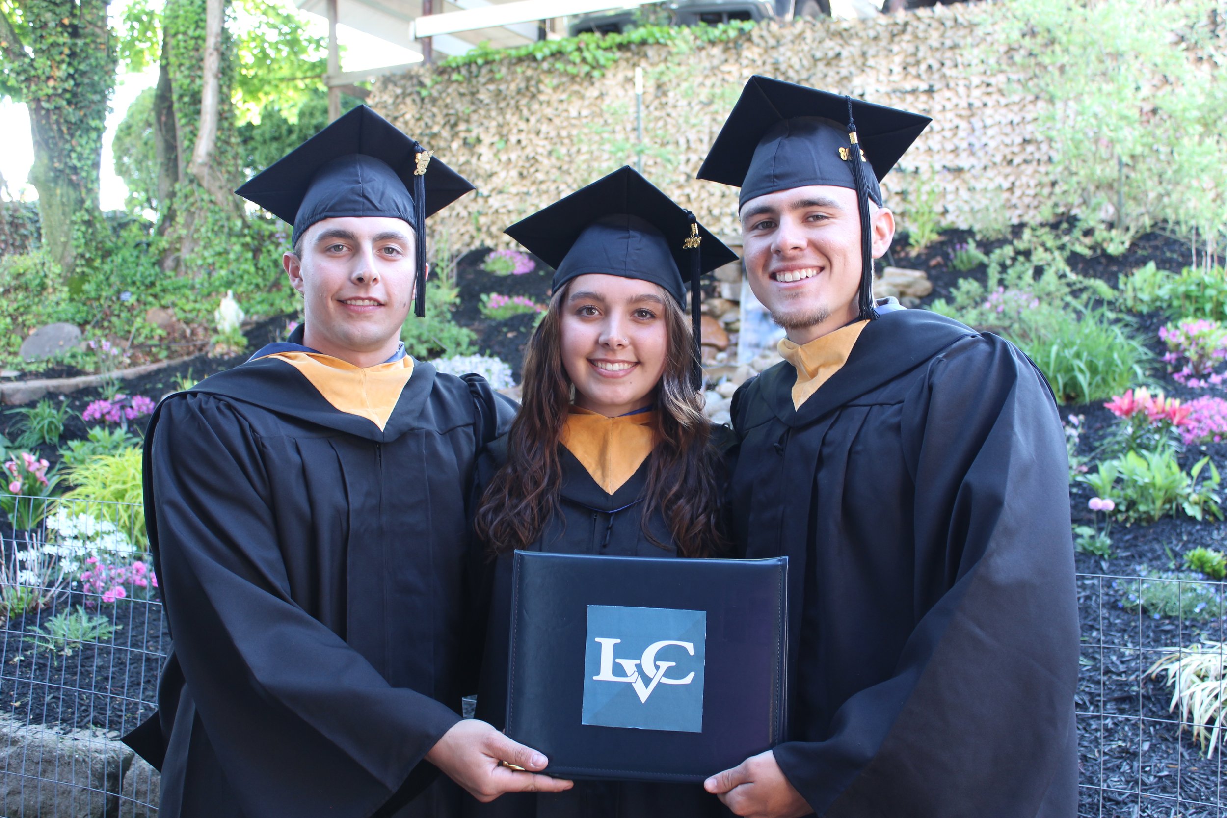 Three young graduates in black caps and gowns holding diploma covers, smiling outdoors in a garden setting.