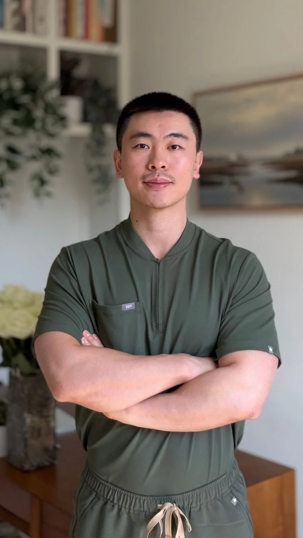 A young Asian man with short black hair, wearing a dark green athletic shirt and matching shorts, stands with arms crossed in a room with a bookshelf, a large painting, and a potted plant in the background.