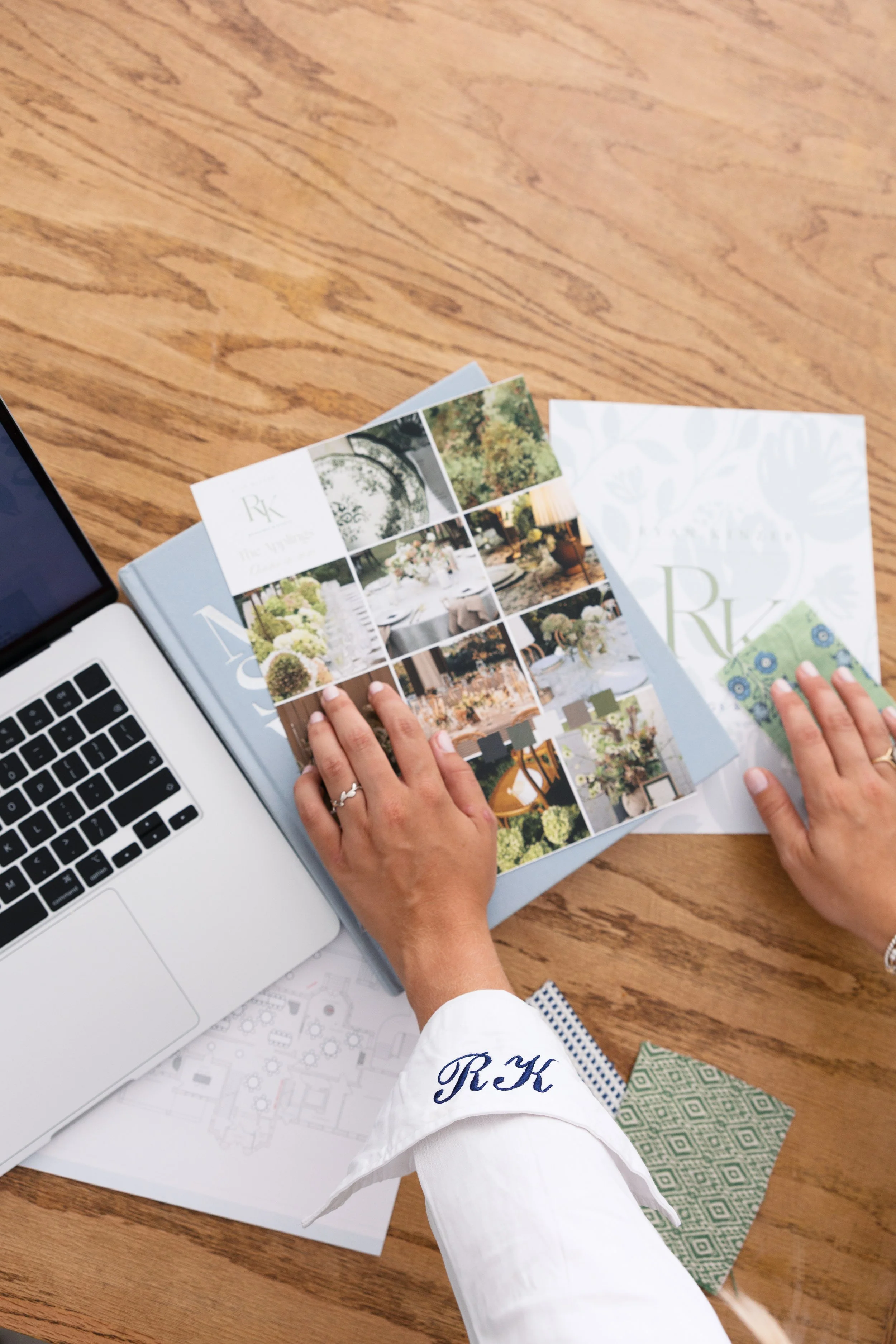 Person holding a colorful brochure or magazine with interior design or event planning images, on a wooden table with a laptop, printed plans, and fabric samples nearby.