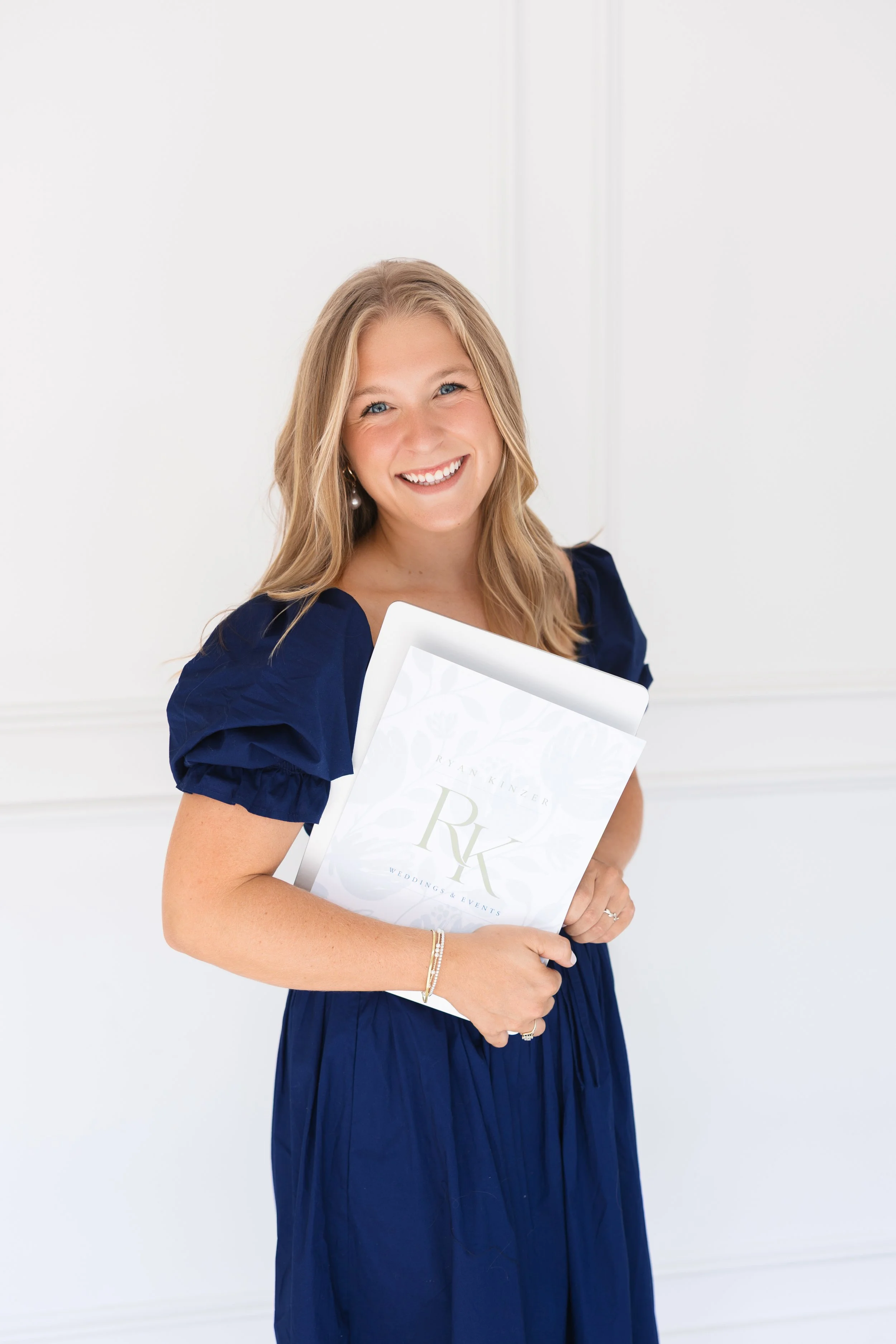 A smiling woman with long blonde hair, wearing a navy blue dress and pearl earrings, holding a white wedding or event planning portfolio.