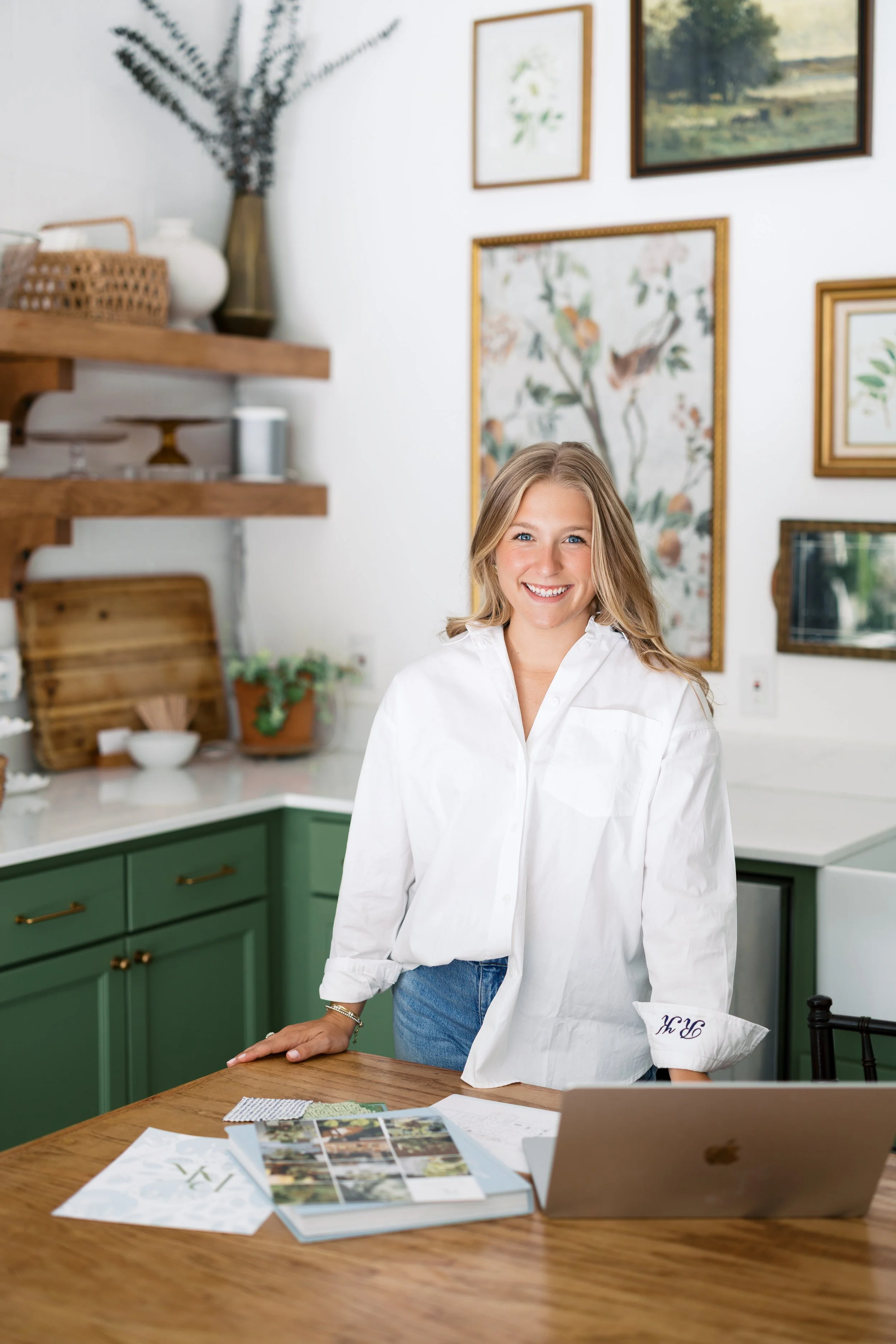 A woman with blonde hair smiling and wearing a white shirt standing in a kitchen with green cabinets and artwork on the wall, with various papers and a laptop on the wooden table in front of her.