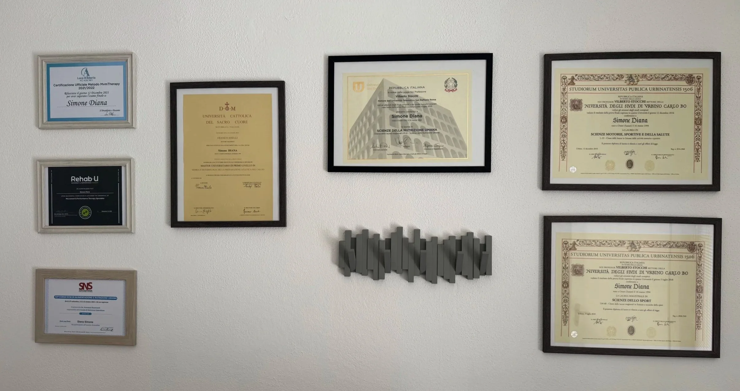 Una parete con diversi diplomi e certificati incorniciati, alcuni in posizioni verticali e altri orizzontali, e una decorazione 3D posta sotto i diplomi.