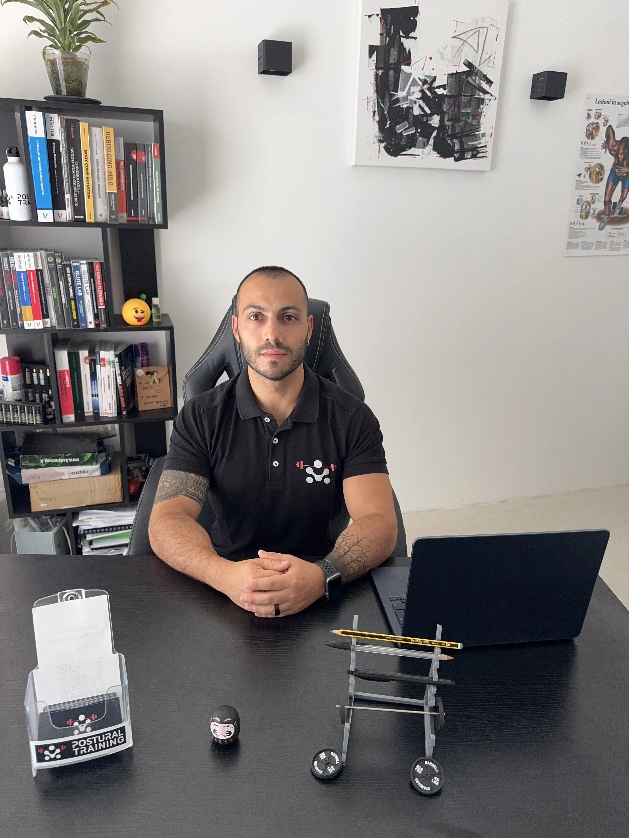 Uomo seduto a un tavolo nell'ufficio, con una libreria piena di libri dietro di lui. In piedi sulla libreria ci sono alcune piante e poster alle pareti. Sulla scrivania ci sono alcuni attrezzi da palestra e un laptop.