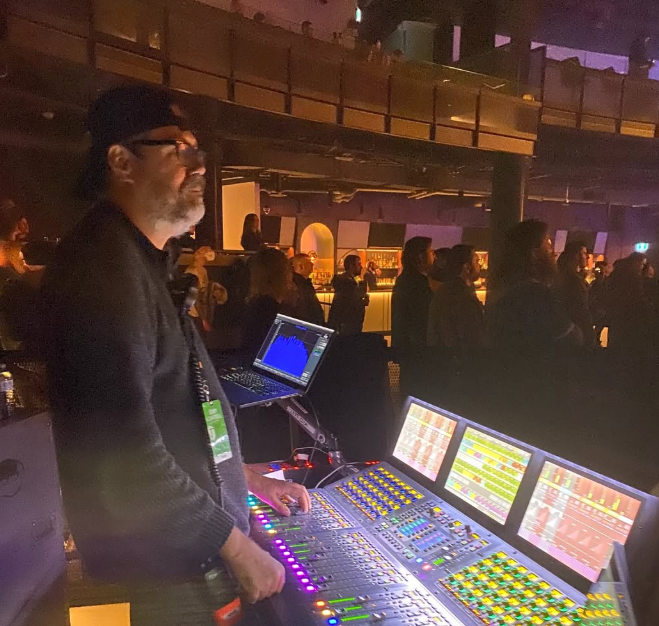 Sound engineer operating a large sound mixing console at an indoor event with people in the background.