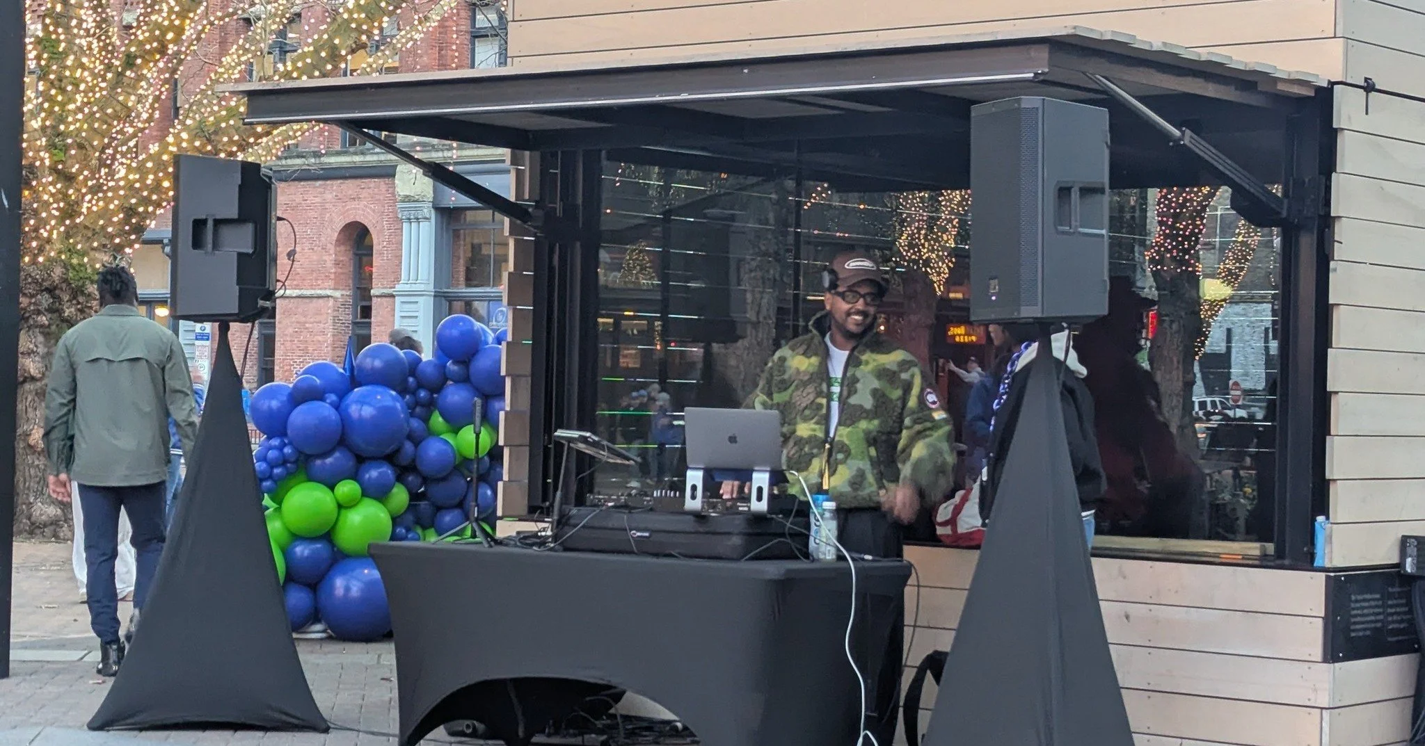 A street scene with a DJ smiling behind a table with electronic equipment in a glass kiosk, decorated with blue and green balloons, and people walking in the background, including a man with headphones and a woman with a backpack. There are trees with string lights and brick buildings in the background.