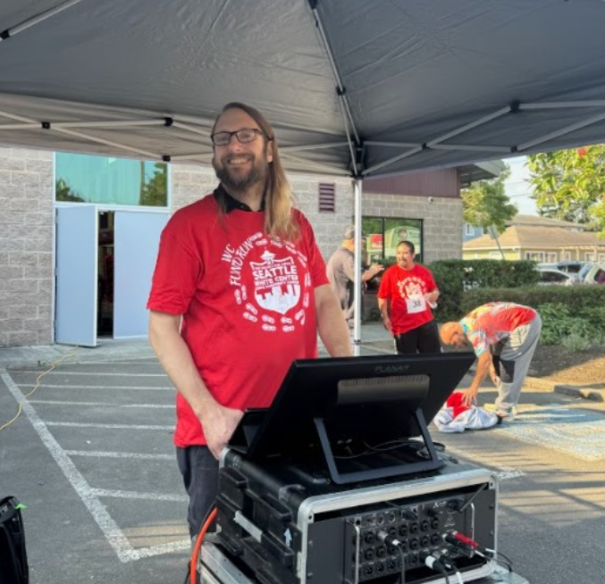 A smiling man with long hair and glasses standing under a canopy near sound equipment, with people in t-shirts in the background preparing for an outdoor event.