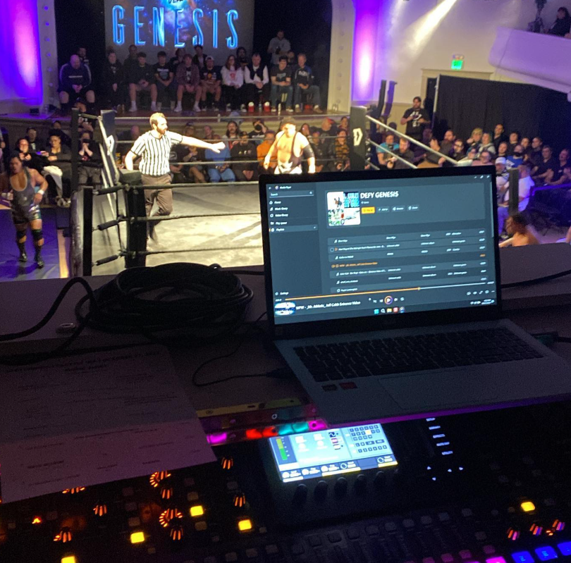 View from a DJ's booth observing a wrestling match in a ring with an audience, stage lighting, and a large screen reading 'GENESIS' in the background.