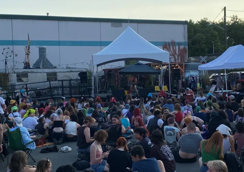 Outdoor concert with a large crowd sitting on the ground, facing a stage with a white canopy, where performers are performing. Tents and equipment are visible behind the stage.
