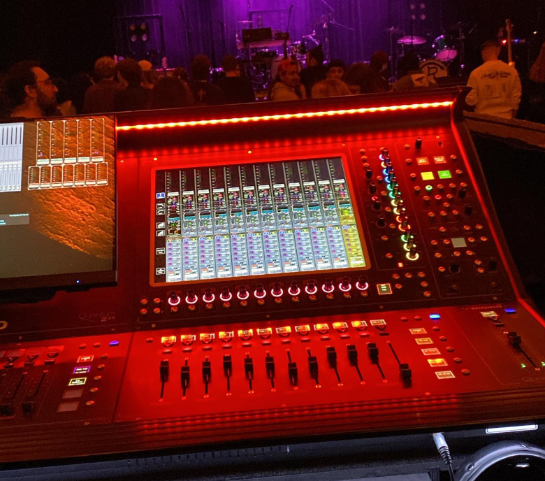 A digital sound mixing console with illuminated controls in a dimly lit venue, with a stage and audience in the background.
