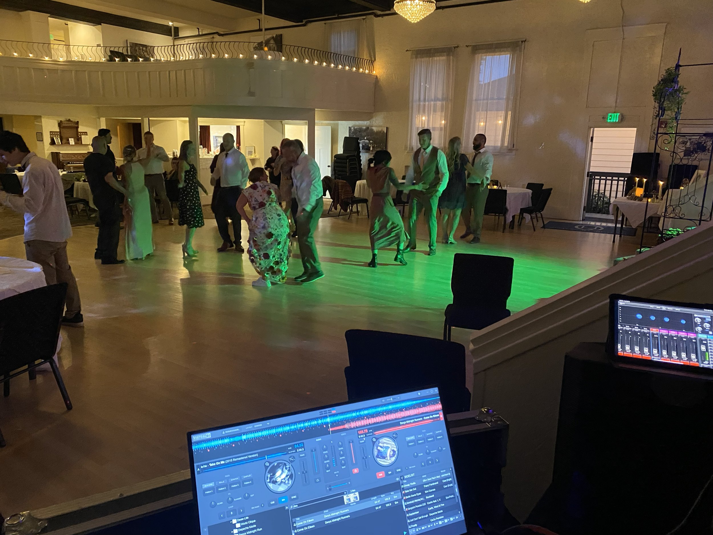 People dancing in a spacious indoor venue with tables, curtains, and a balcony. A DJ setup with music software is visible in the foreground.