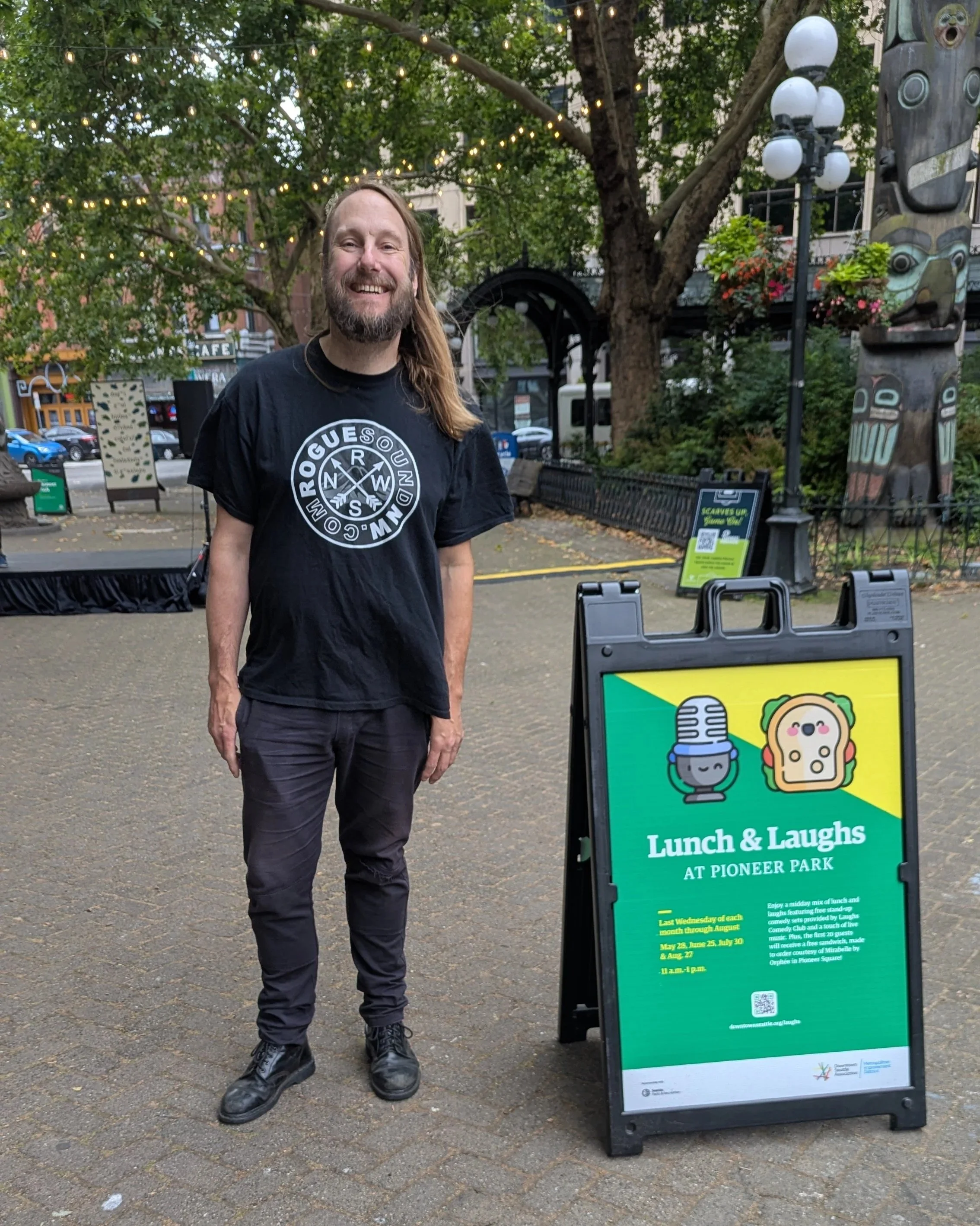 A man with long hair and a beard, wearing a black T-shirt with the word 'GUESS' and a graphic design, standing outdoors next to a sign that promotes Lunch & Laughs at Pioneer Park. The sign has colorful graphics and text mentioning dates and details about the event. Behind him are trees, string lights, a building, and decorations including totem poles.