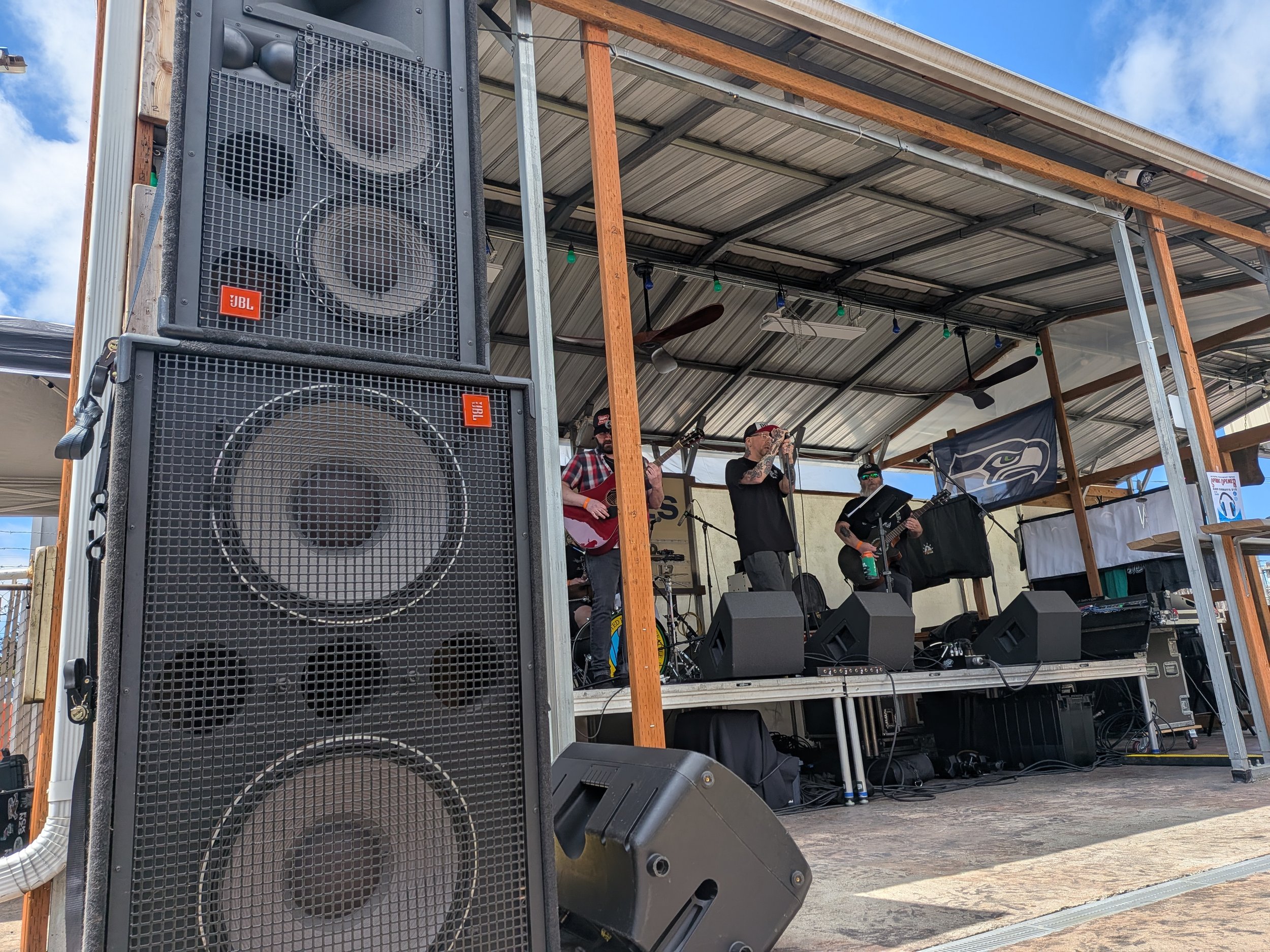 A stage with musicians performing, a large JBL speaker in front, and a metal roof structure with string lights above. The stage is outdoors with a Seattle Seahawks banner hanging and a partly cloudy sky.