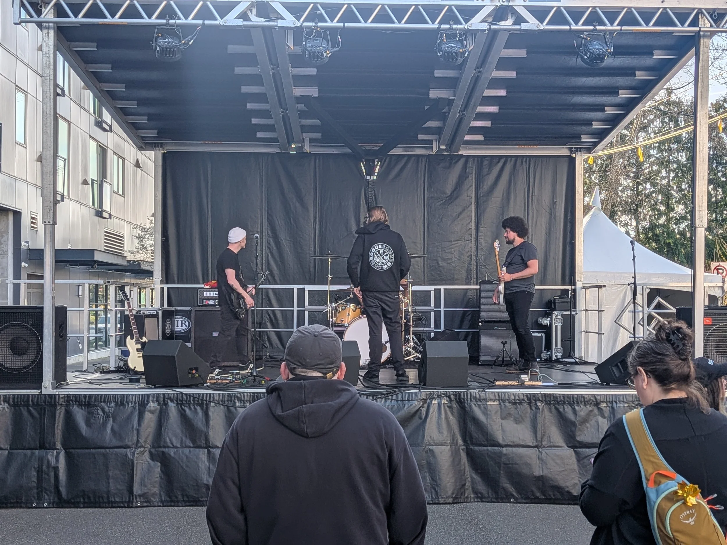 A stage set up outdoors with three men preparing for a performance. One man is holding a guitar, another is facing away wearing a hoodie with a band logo, and the third is standing with a guitar. Audience members are visible in the foreground.