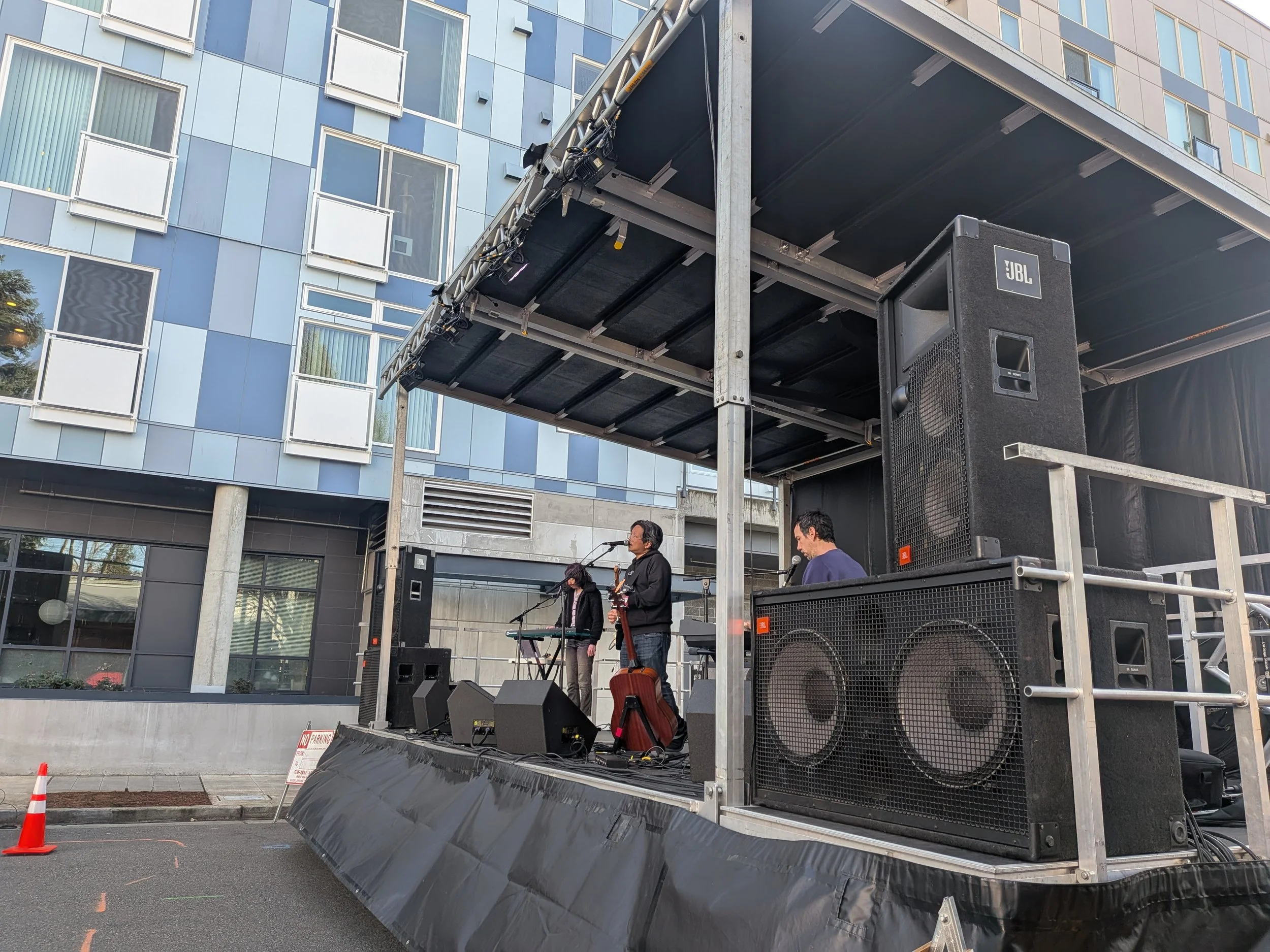 A stage set up outside in front of a modern glass building with microphones and musical instruments, where a band is performing.