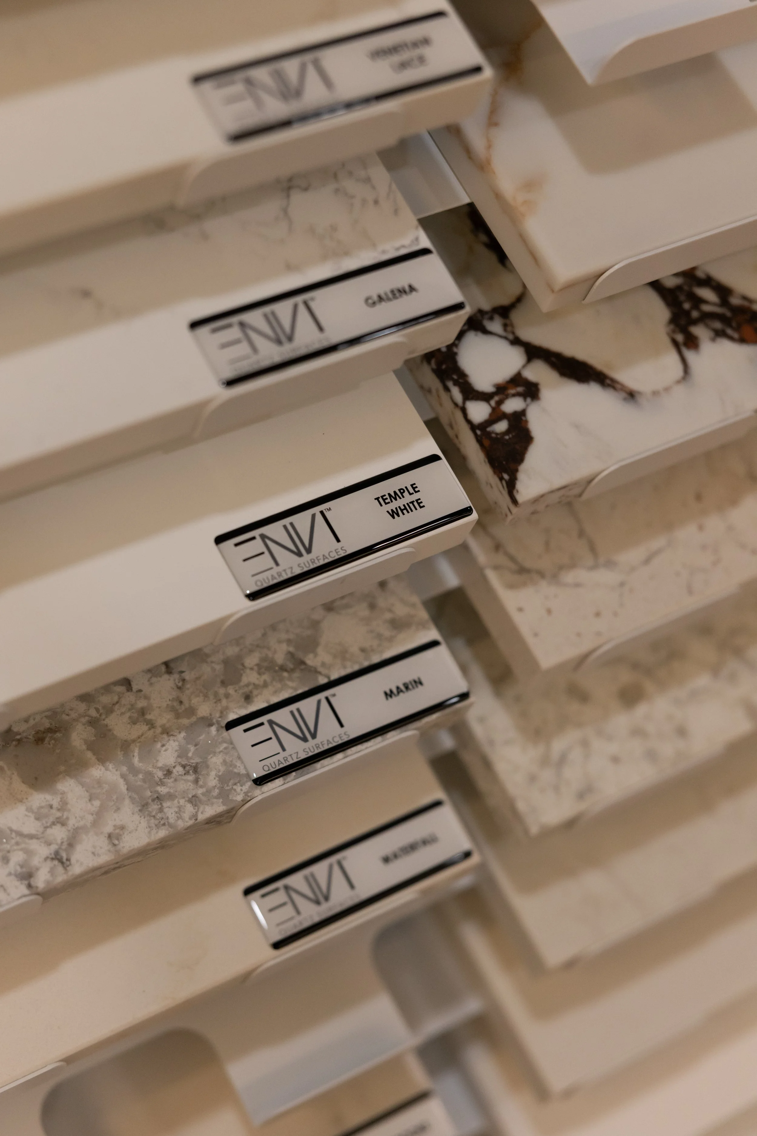 Close-up of color samples, including white marble, with labeled tags such as 'Tangent', 'Galena', 'Temple White', and 'Marin', arranged on shelves.