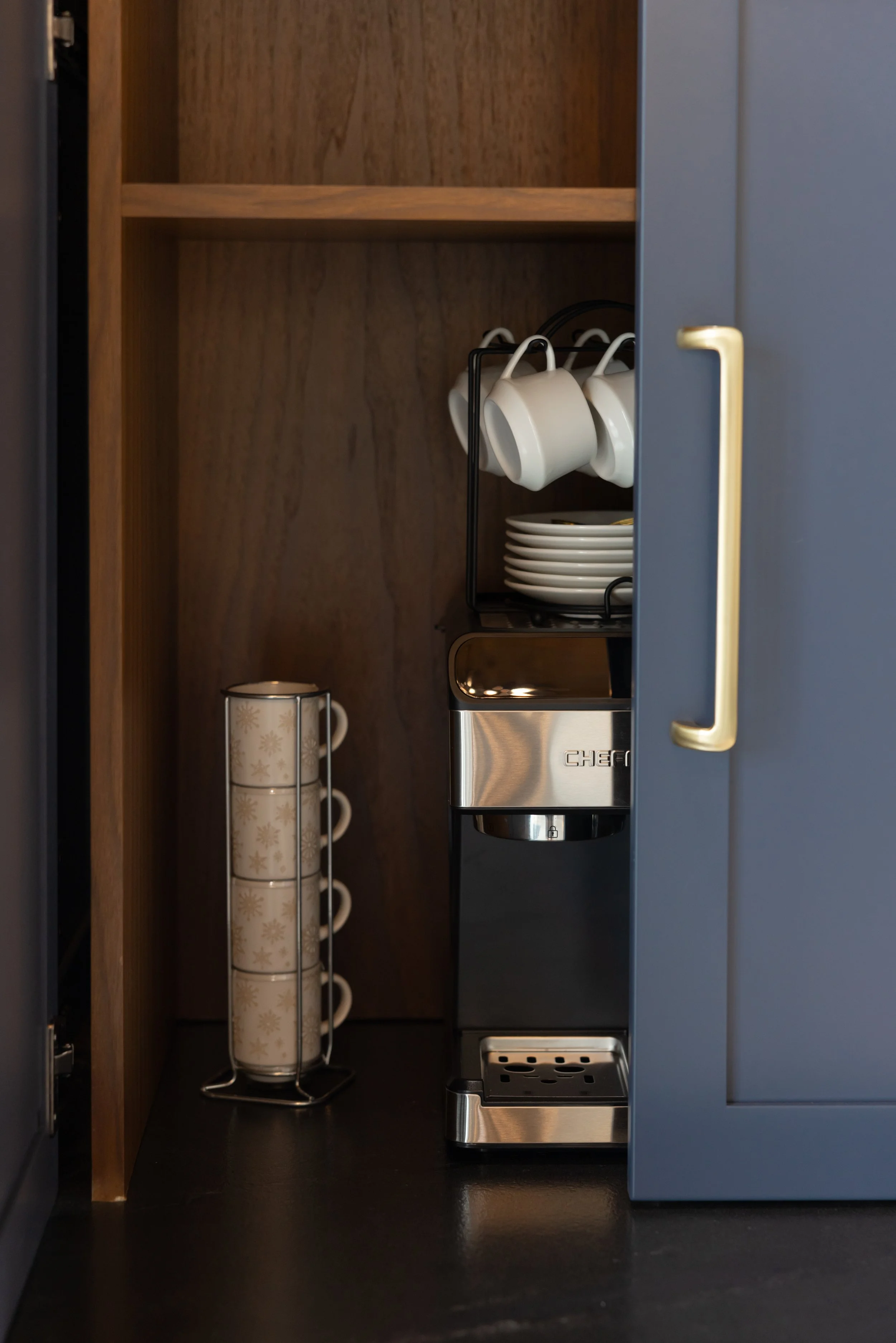 Inside a kitchen cabinet with a coffee machine, white mugs hanging on hooks, a stack of small plates, and a mug with a snowflake pattern on a mug tree.