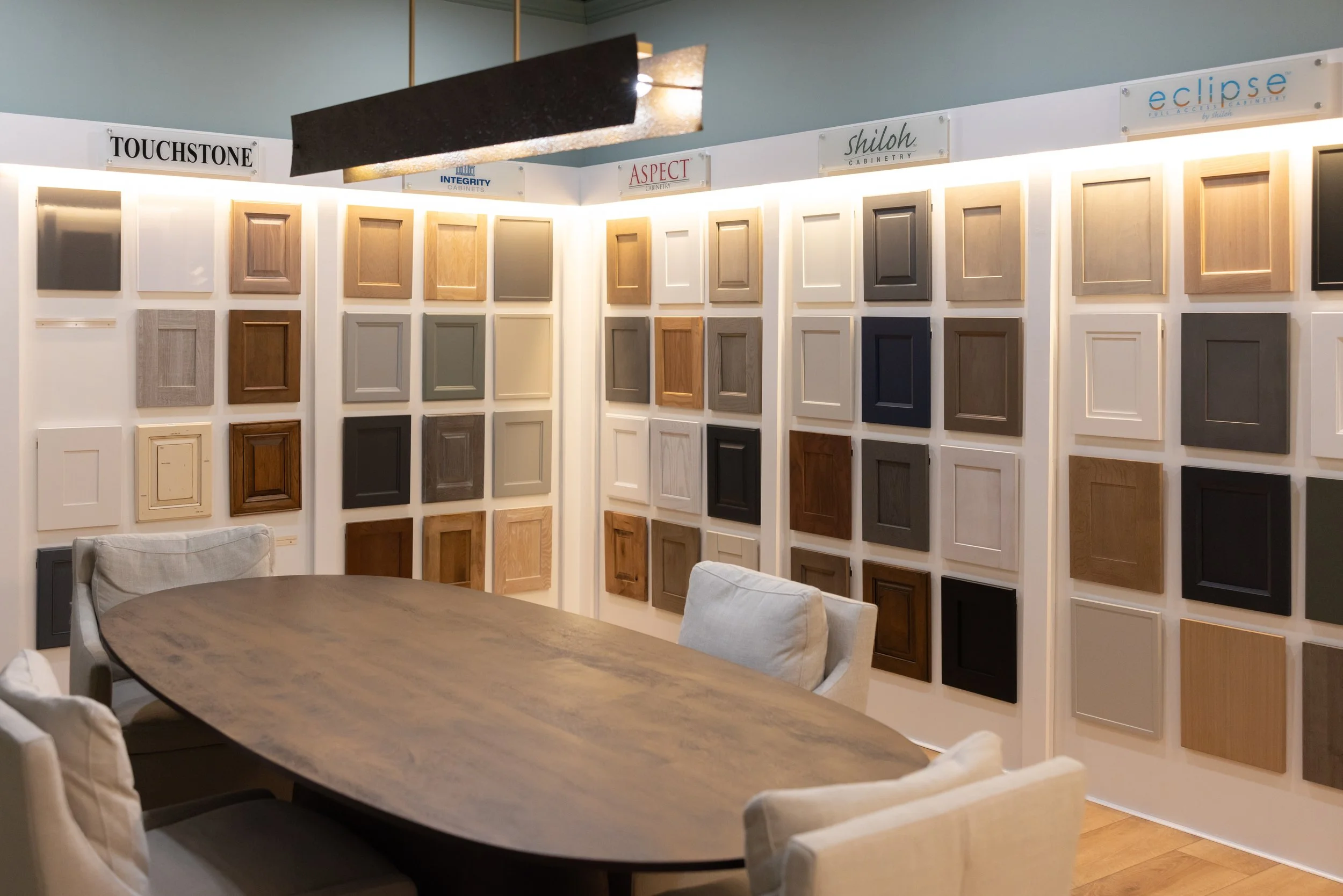 Display of various cabinet door samples on a wall in a showroom with a wooden table and chairs in the foreground.