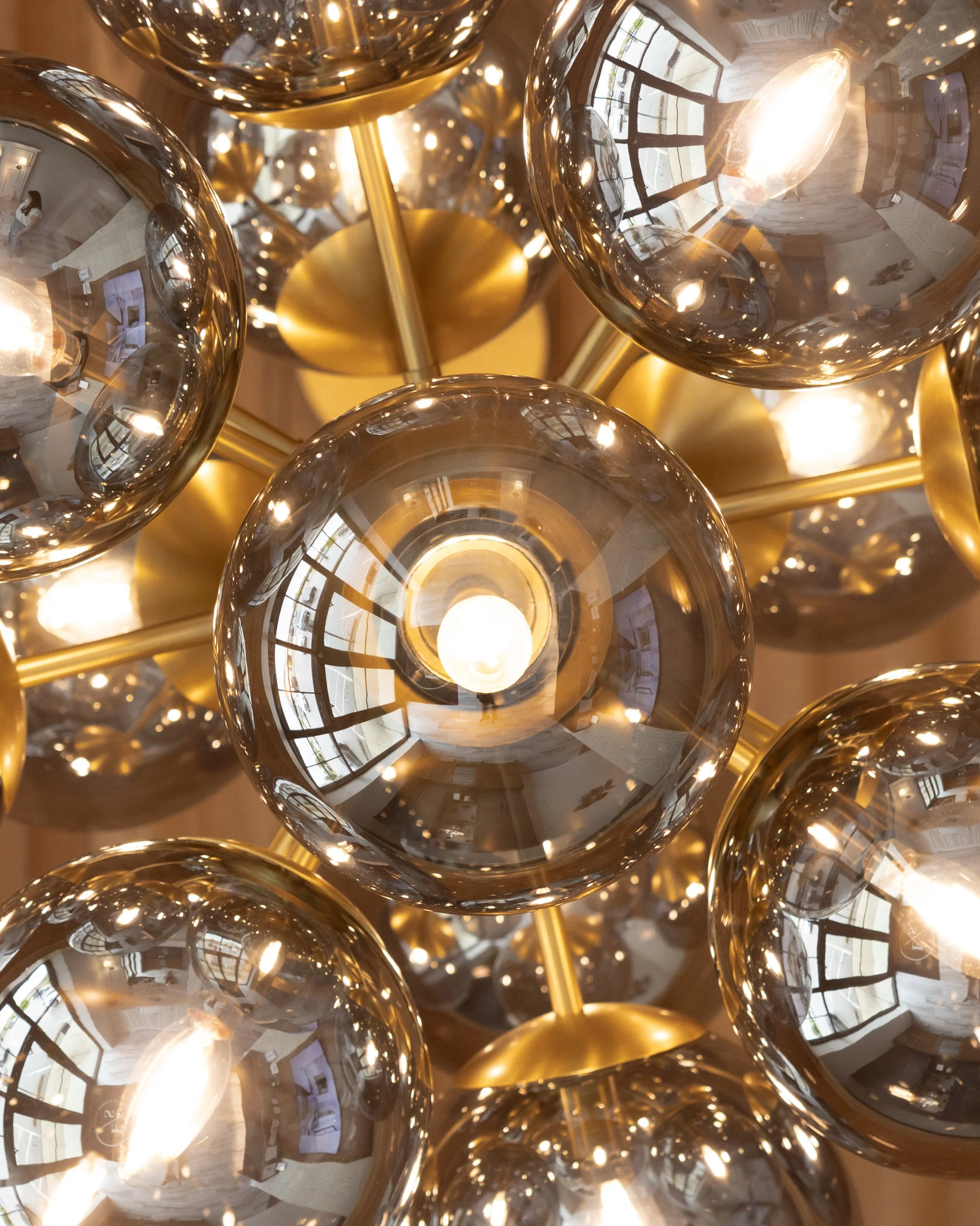 Close-up view of a modern chandelier with multiple reflective, gold-colored spherical glass globes, reflecting interior rooms including windows, doors, and furniture.