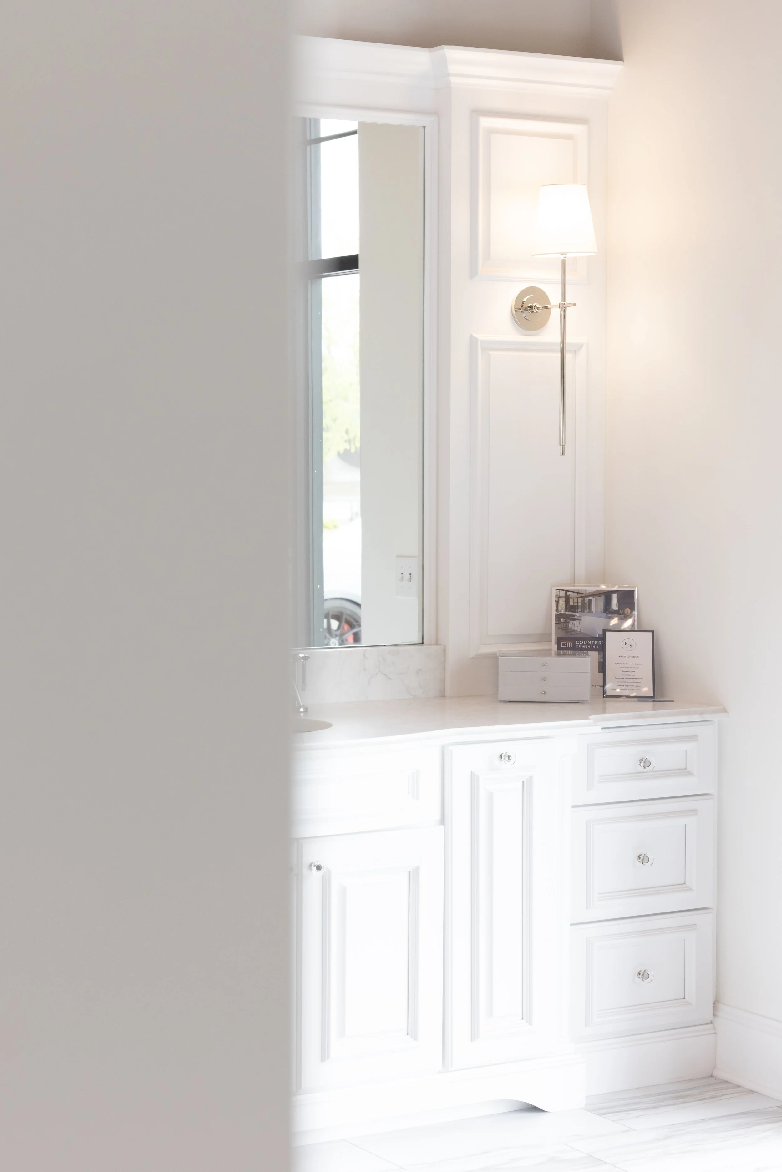 A white bathroom vanity with four drawers and cabinets, a marble countertop, a tall mirror, a window, a wall-mounted lamp, and framed pictures on the counter.