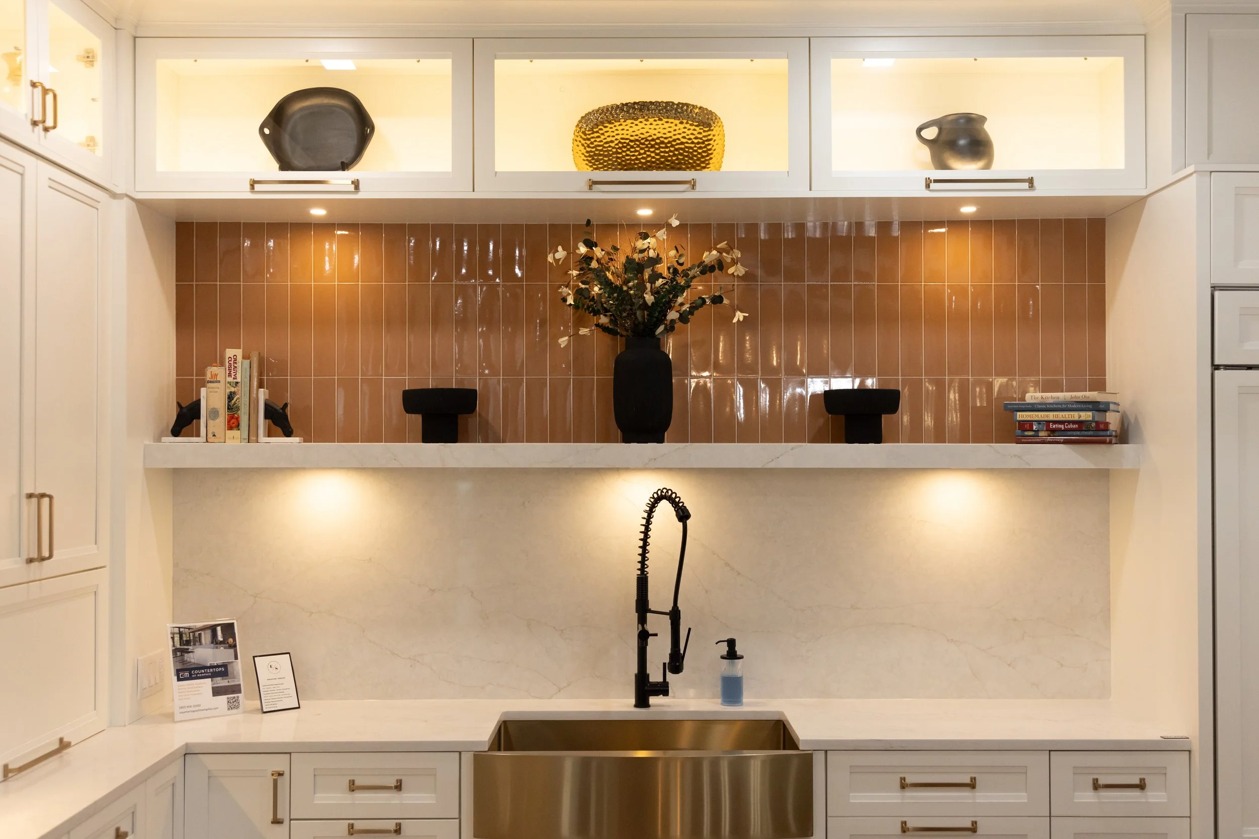 Modern kitchen with white cabinets, brown tiled backsplash, marble countertop, center sink, black faucet, and decorative plates and a vase with flowers on the backsplash