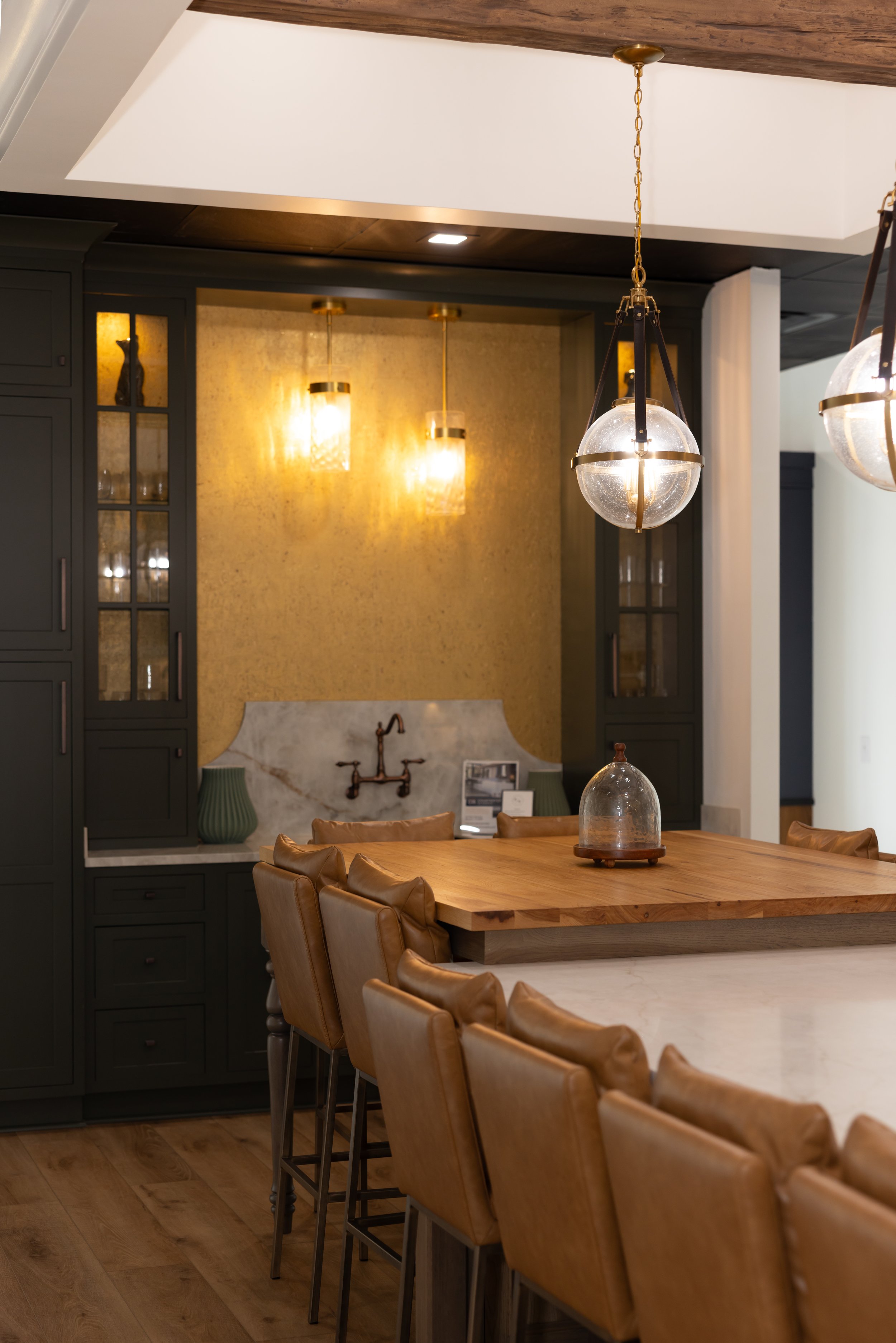 Inside a modern dining area with a wooden table and tan leather chairs. Hanging light fixtures with glass globes illuminate a dark cabinet with glass-paneled doors and decorative items behind the glass. The background features a gold-toned wall and a marble countertop with a decorative faucet.