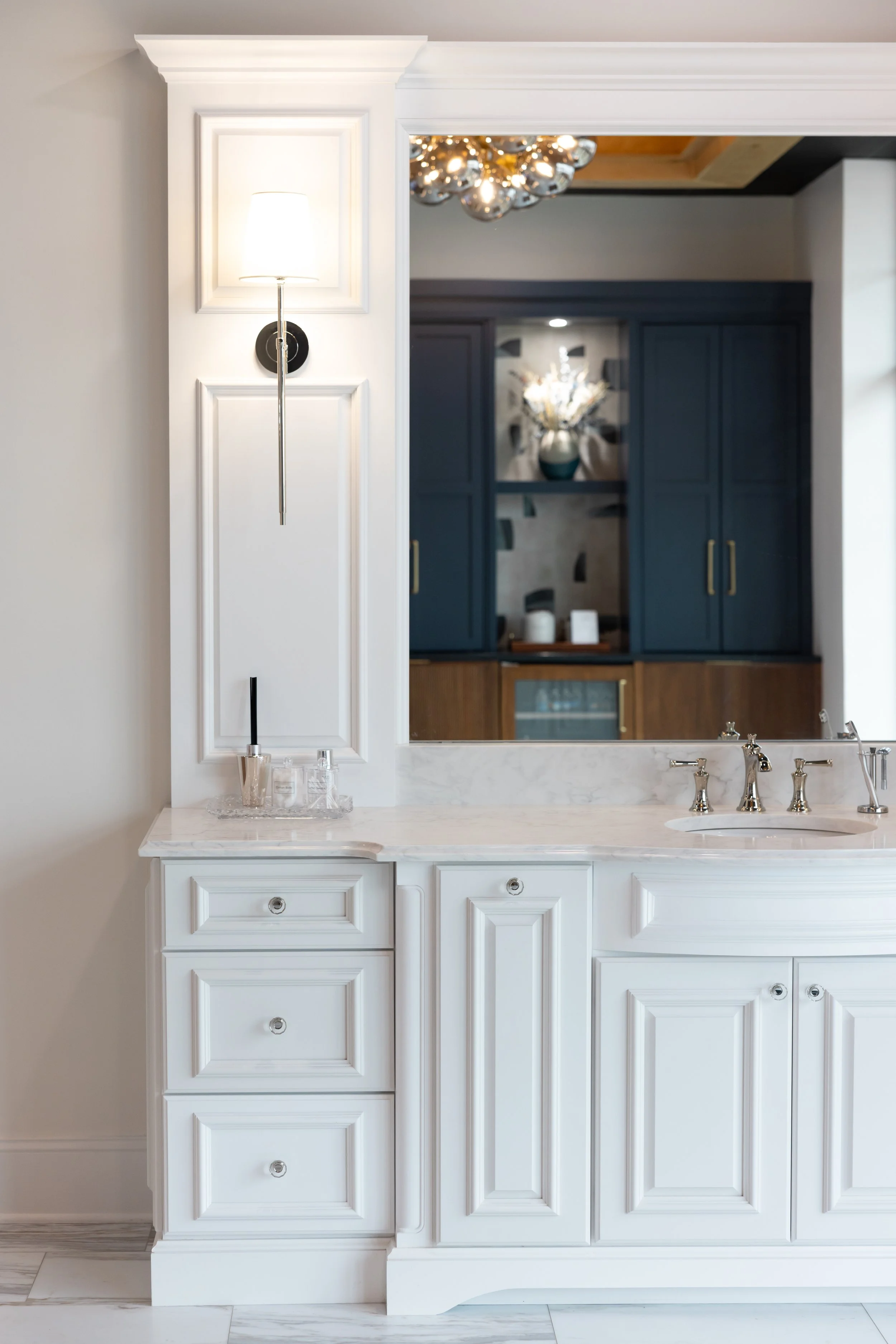 Modern bathroom vanity with white cabinetry, marble countertop, and gold fixtures. A large mirror reflects a blue cabinet and a decorative vase with flowers.