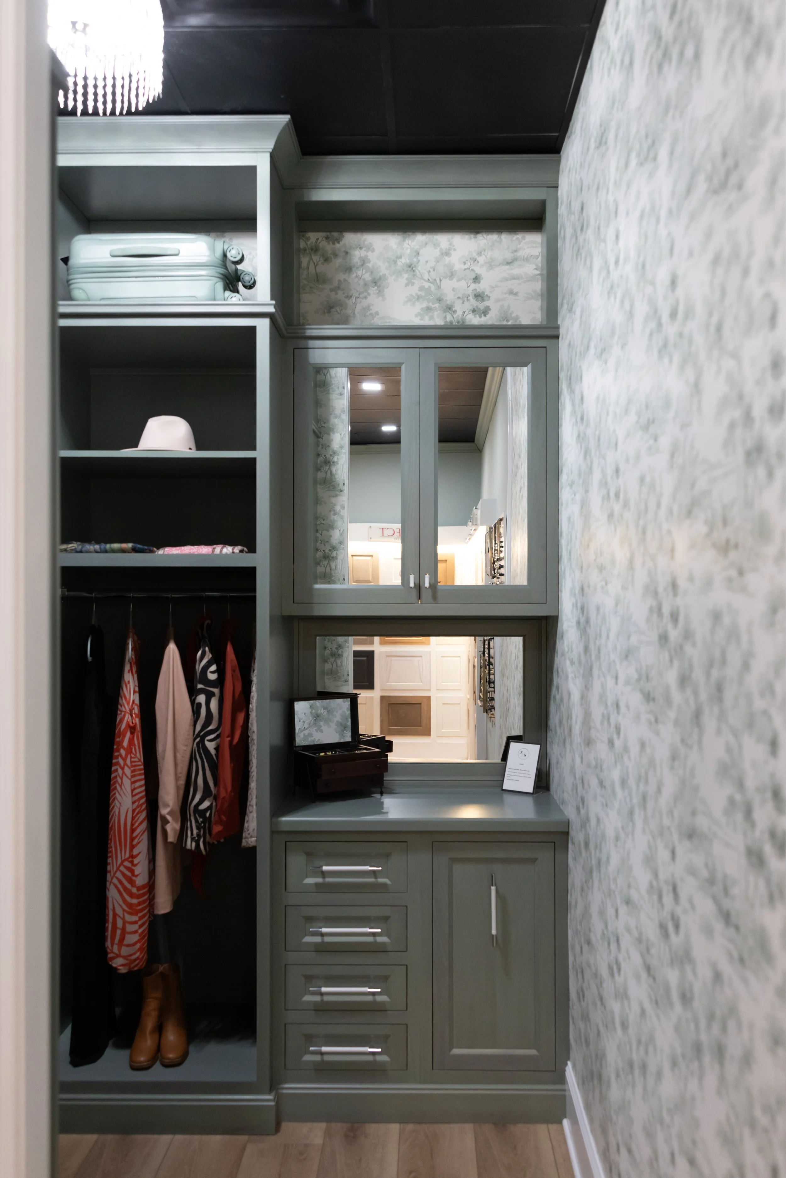 A built-in greenish-gray cabinet with glass-paneled doors, drawers, and open shelves, situated within a hallway with floral-patterned wallpaper. The shelving holds a white plastic pair of shoes on the top left, and a white lamp on the middle shelf. A mirror at the bottom reflects interior room details, including framed images and part of a ceiling with recessed lighting.