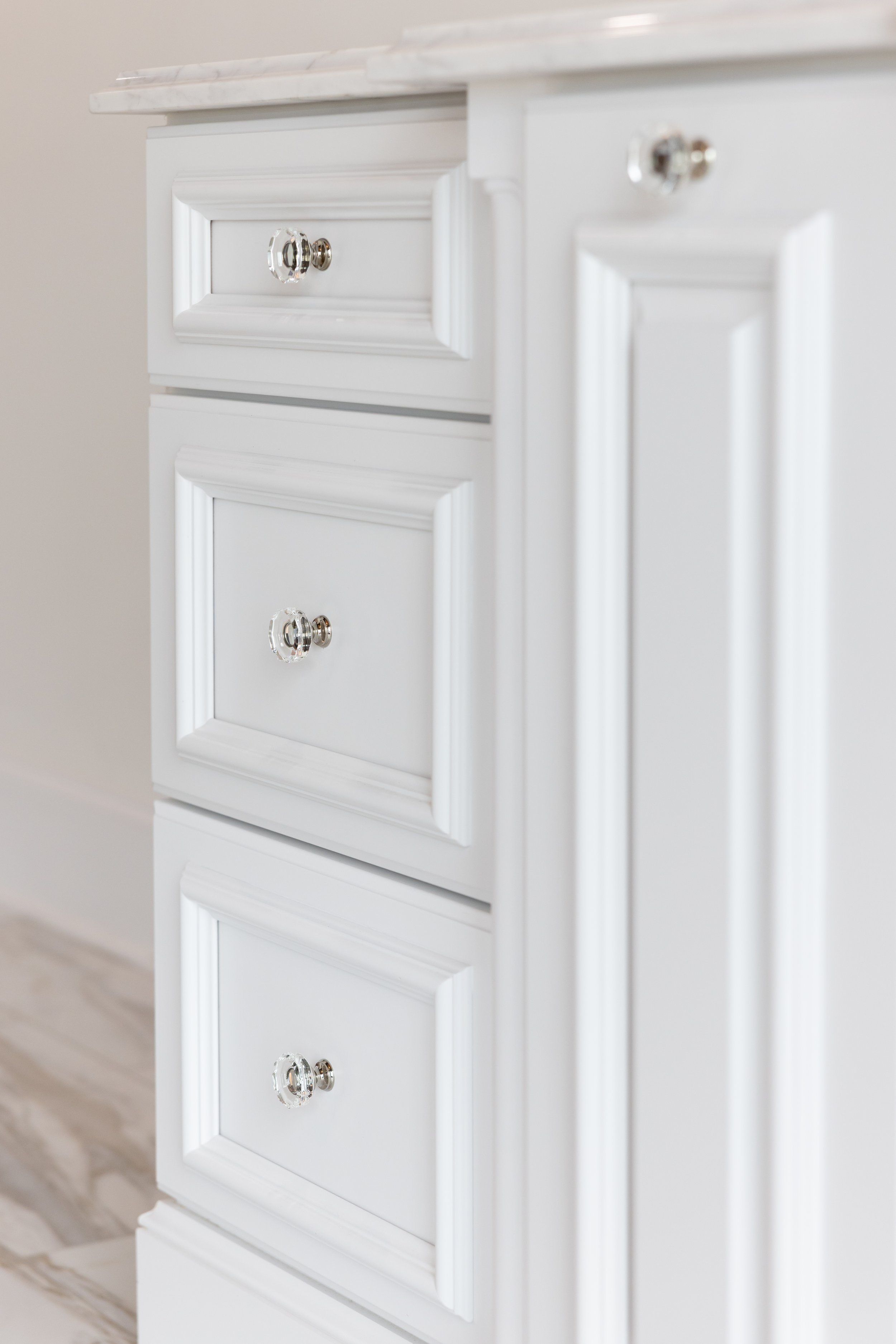 Close-up of a white wooden cabinet with silver and clear crystal knobs, three drawers visible, with a white marble top.