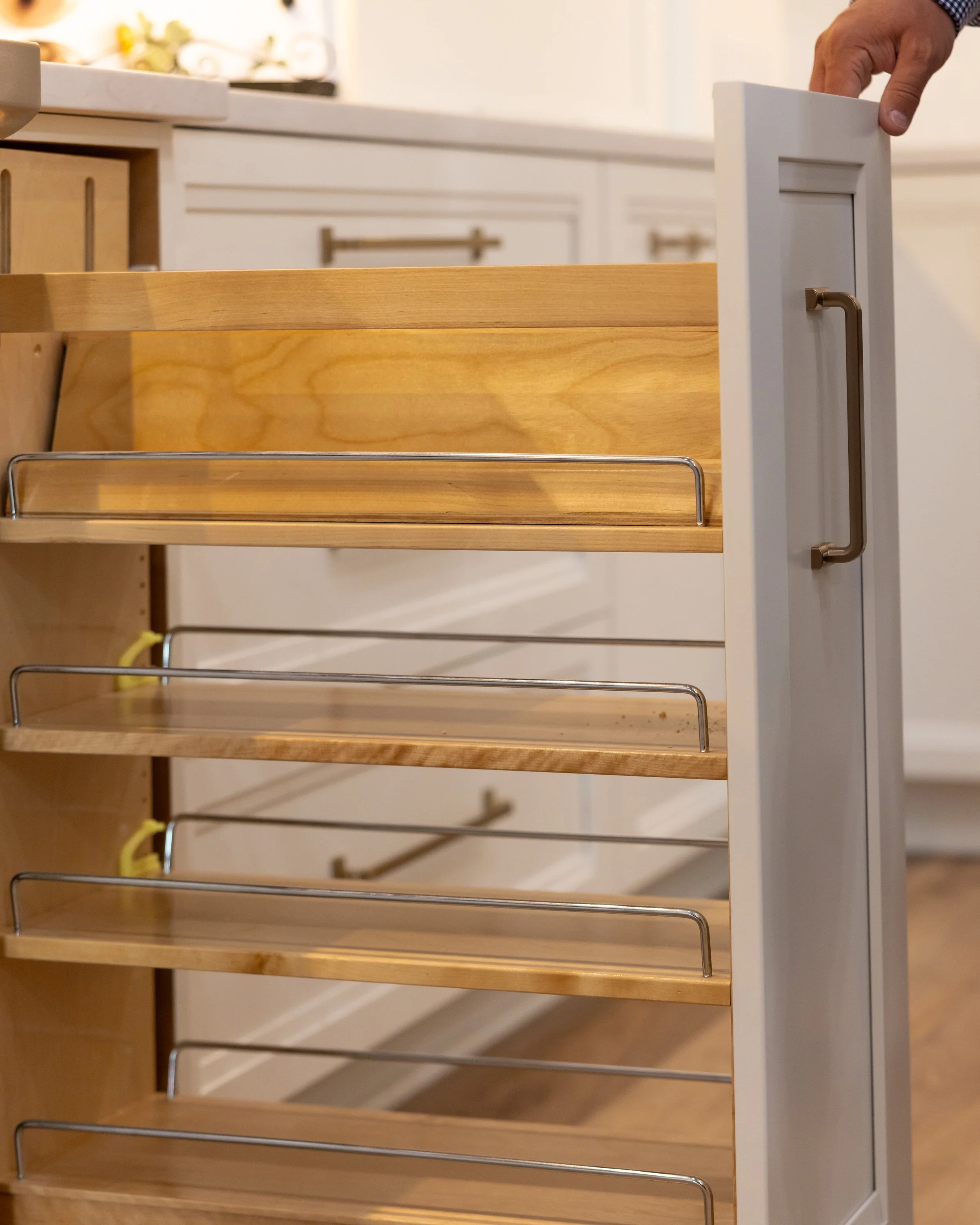 A person opening a white cabinet drawer in a kitchen, with wooden shelves and a countertop with flowers in the background.