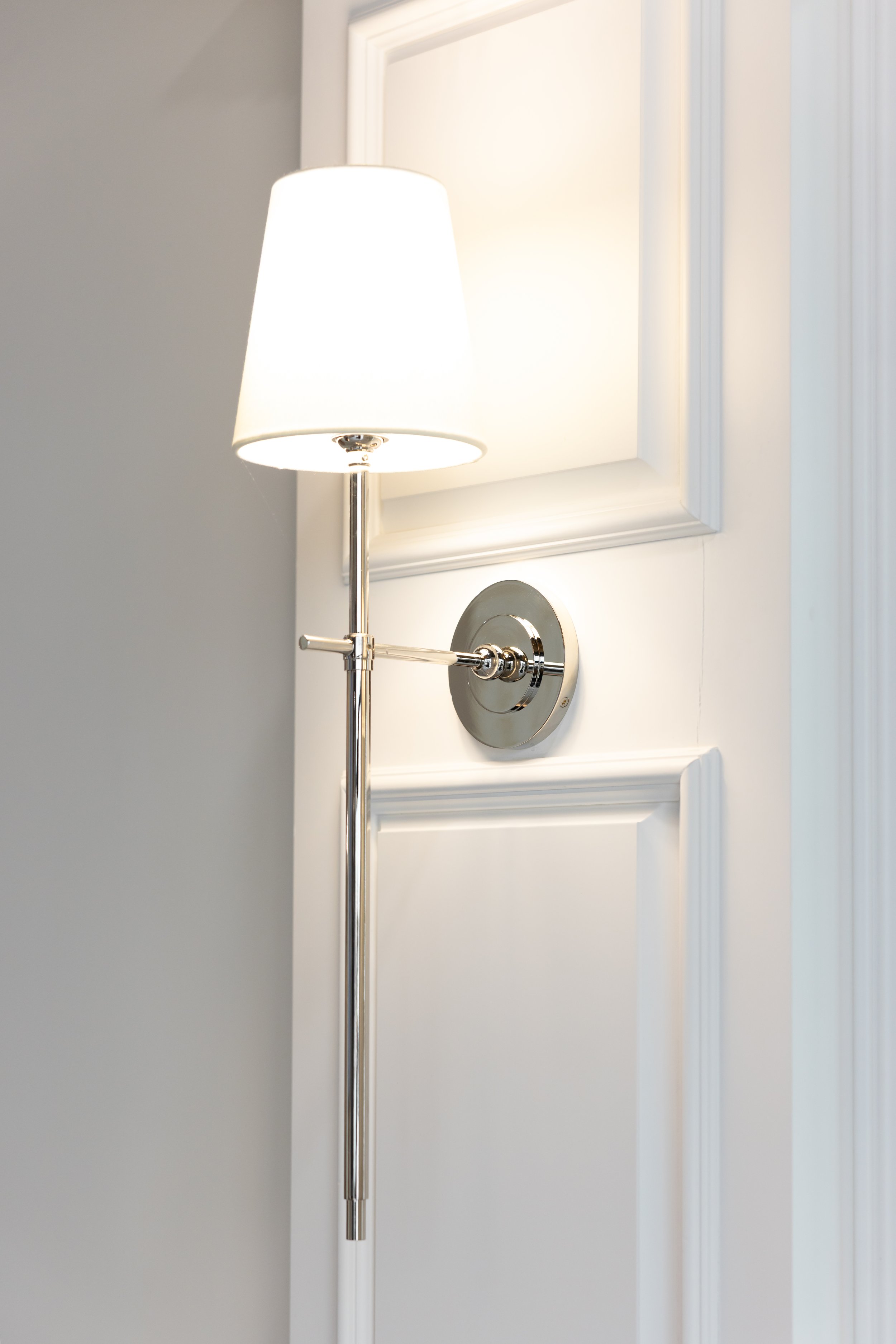 A white wall-mounted lamp with a white lampshade turned on, casting light on a white wall and doorframe.