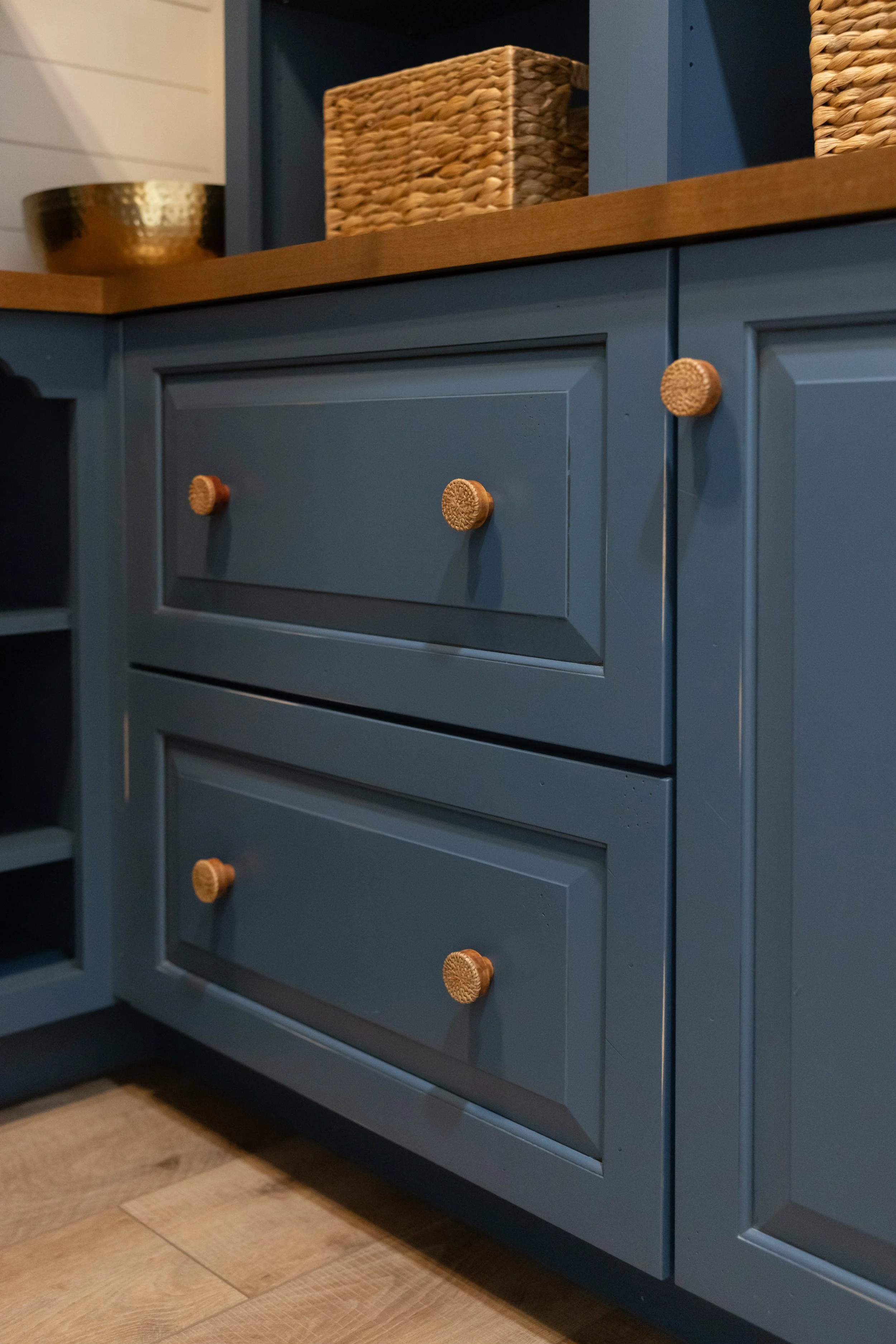 Blue kitchen cabinet with wooden knobs, a drawer, and a wooden countertop. Wicker baskets and a metallic bowl are placed on top of the cabinet. Part of a patterned rug and wooden floor are visible.