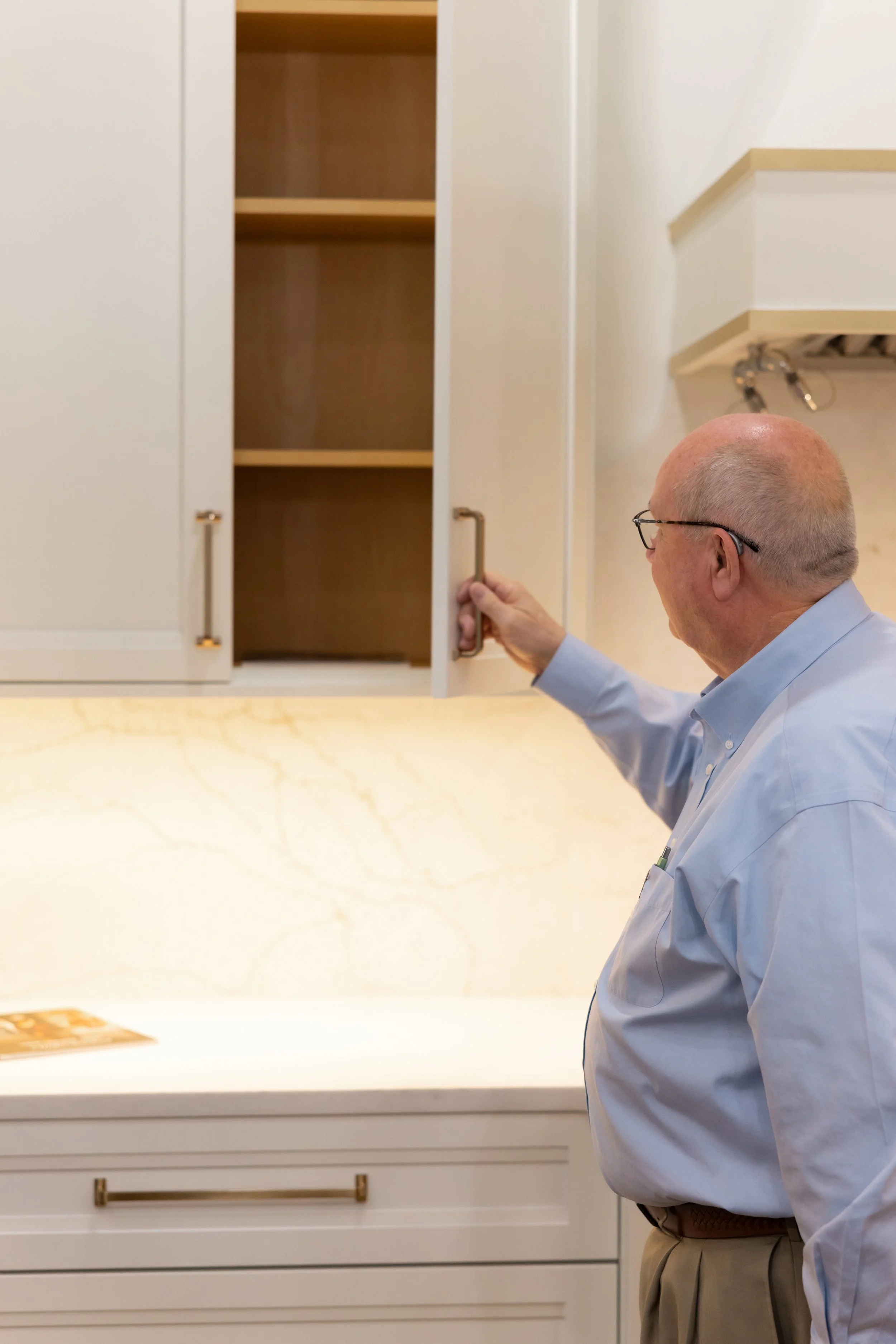 A man with glasses, a light blue shirt, and khaki pants opening a cabinet door in a kitchen with white cabinets and a marble countertop.