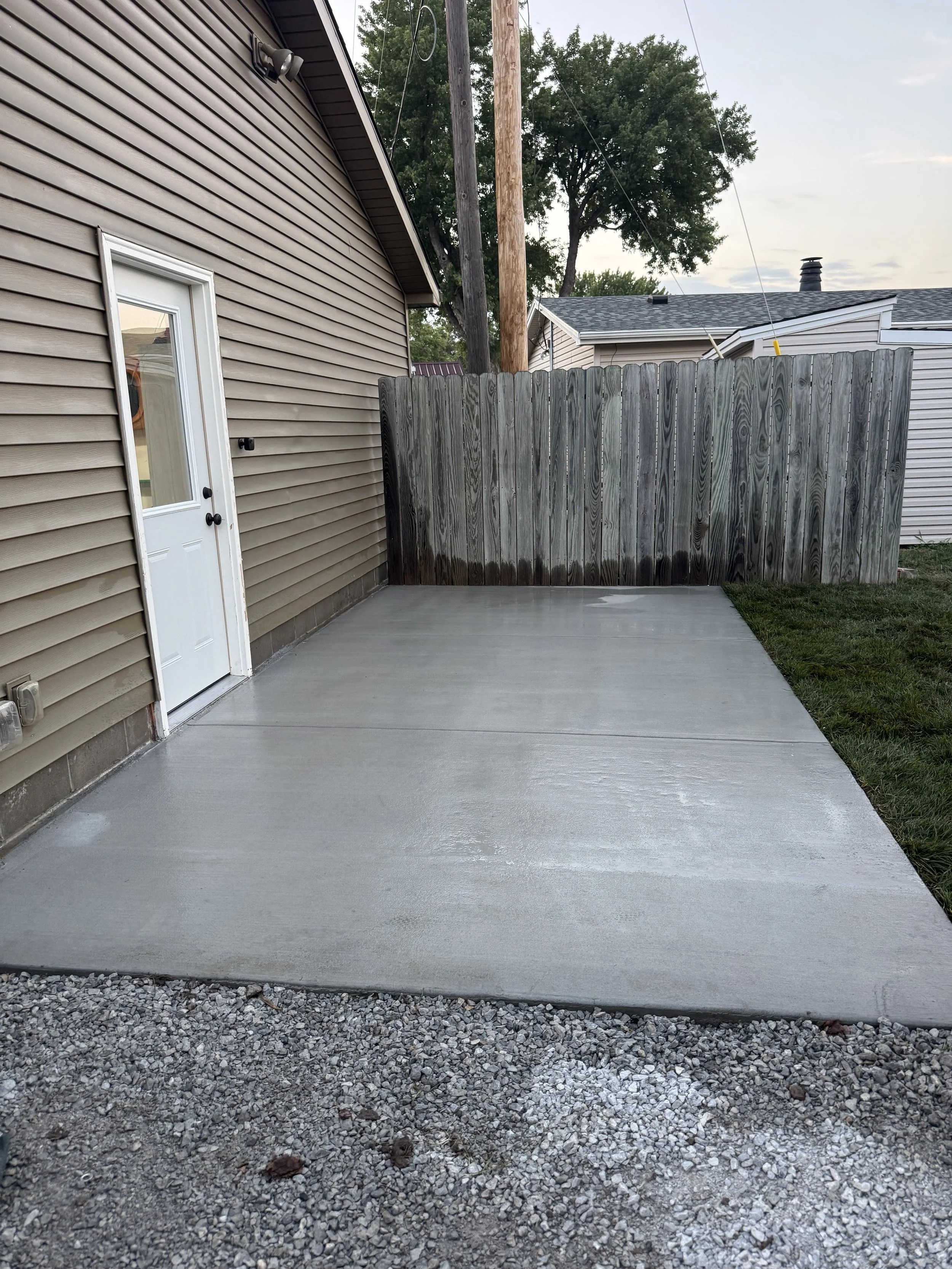 A concrete patio next to a beige house with horizontal siding, a white door, and a black doorknob. To the right, there is a grassy yard with a gravel border. There is a wooden privacy fence at the back with a large tree behind it. The sky is cloudy.