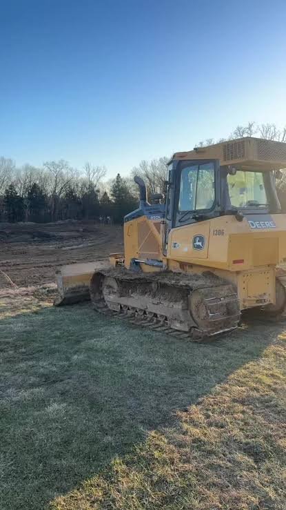 A yellow John Deere bulldozer on a construction site with bare trees and a clear blue sky in the background.