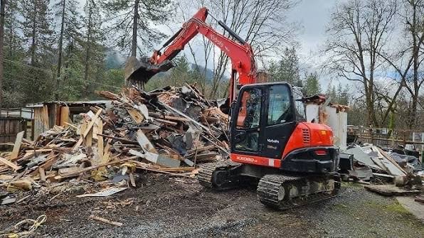 A red compact excavator demolishing a wooden structure in an outdoor setting with trees and a cloudy sky in the background.