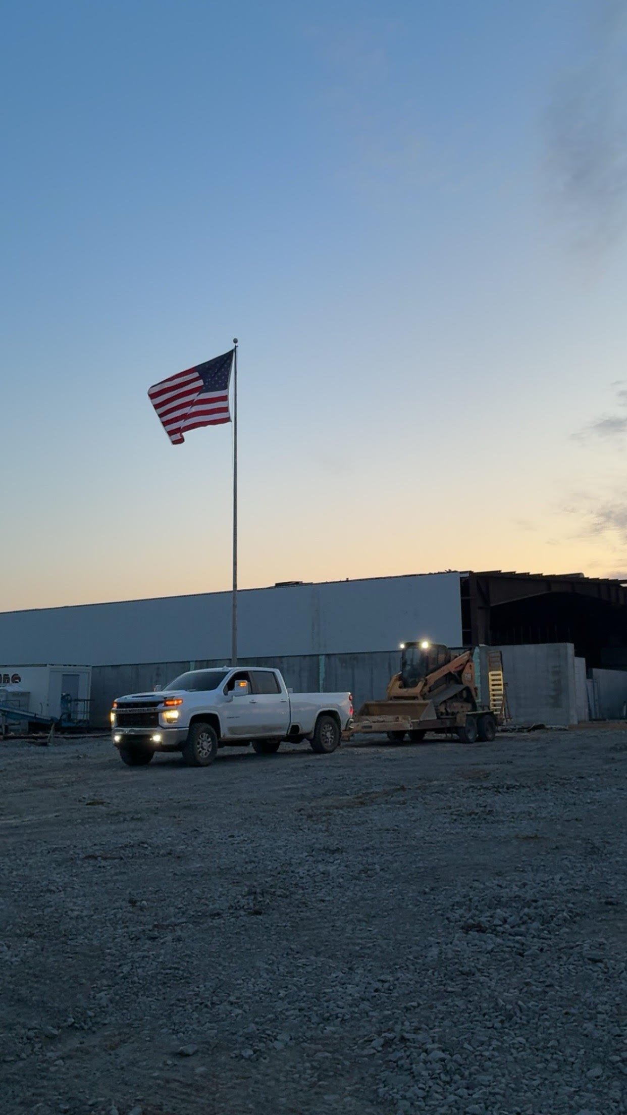 A parking lot with a white pickup truck, construction equipment, and a large American flag flying against a clear evening sky near an industrial building.