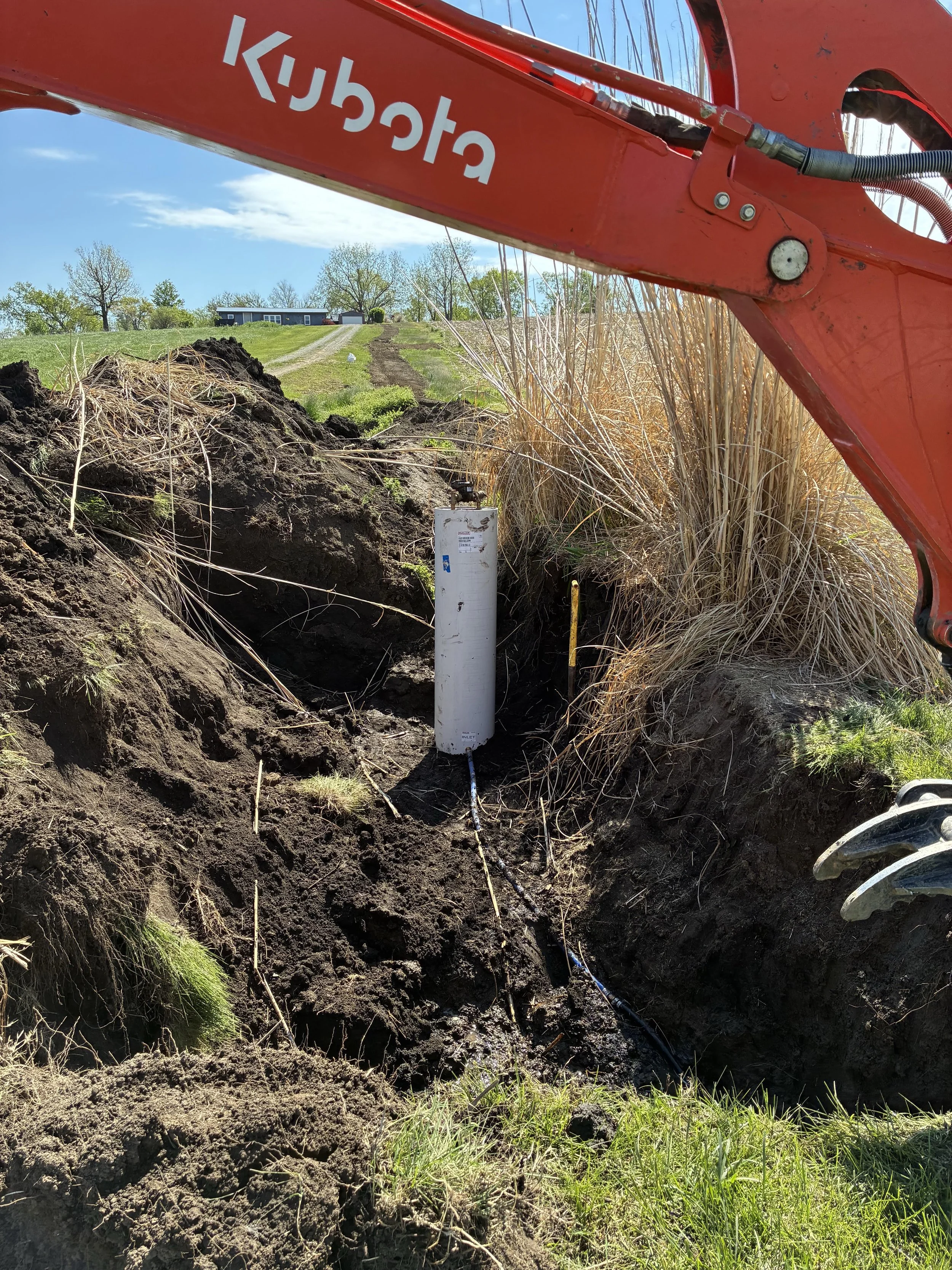 View of a construction site with a large red excavator partially visible in the foreground. There is a deep trench in the earth with some piping and underground utilities visible. A white pipe stands upright in the ground with some dirt and grass surrounding it. In the background, there is a grassy hill with a house, trees, and a gravel driveway under a blue sky with some clouds.
