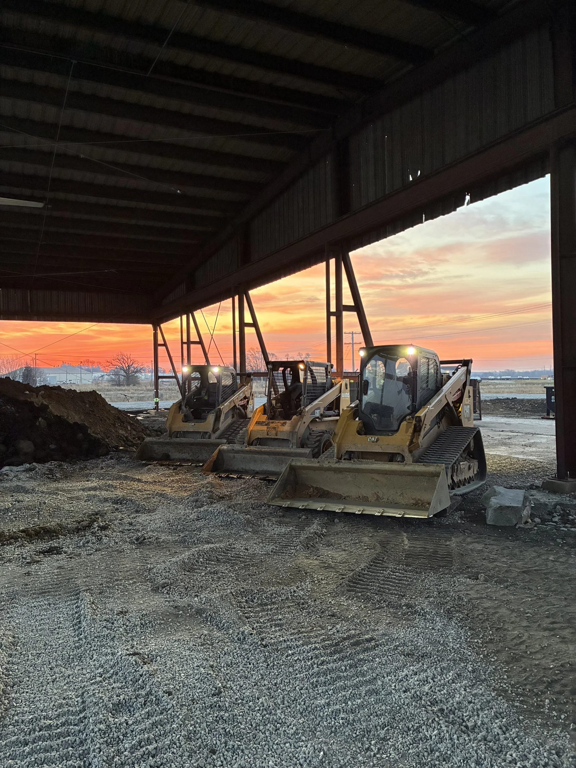 Construction site under a large metal roof with three front loaders parked on gravel, sunset in the background.